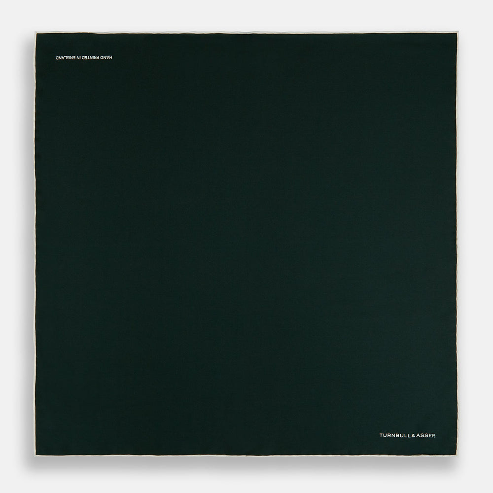 The DARK GREEN AND ECRU PIPED SILK POCKET SQUARE by Turnbull & Asser features ecru edging and the brand's logo in one corner, making it a refined choice for formal attire. One corner is folded to show the reverse side.