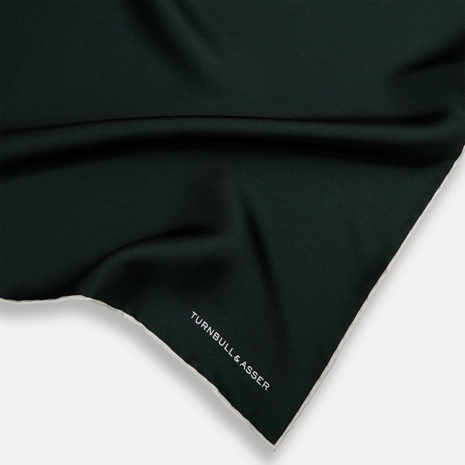 View 2 of product The DARK GREEN AND ECRU PIPED SILK POCKET SQUARE by Turnbull & Asser features ecru edging and the brand's logo in one corner, making it a refined choice for formal attire. One corner is folded to show the reverse side.