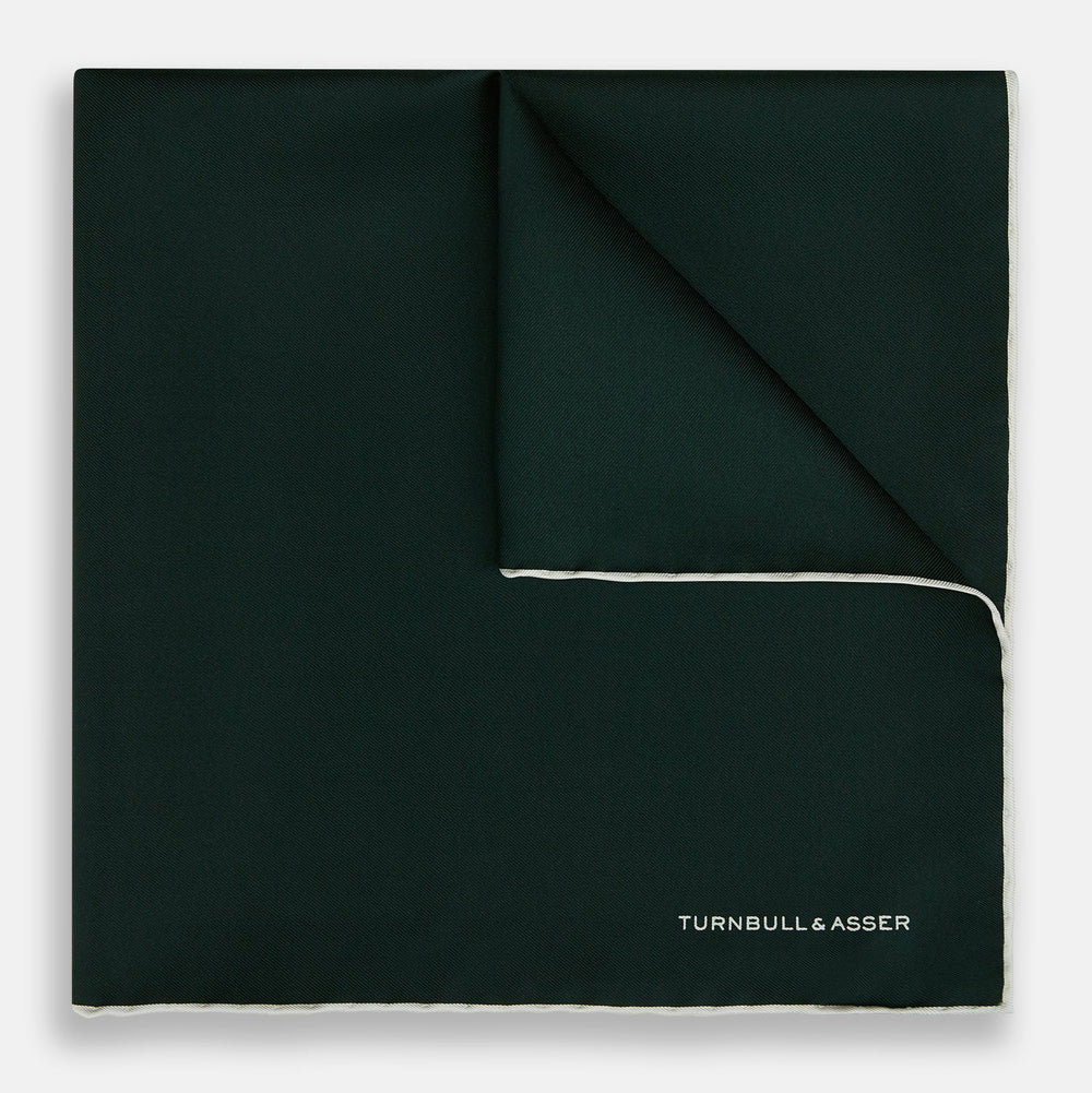 The DARK GREEN AND ECRU PIPED SILK POCKET SQUARE by Turnbull & Asser features ecru edging and the brand's logo in one corner, making it a refined choice for formal attire. One corner is folded to show the reverse side.