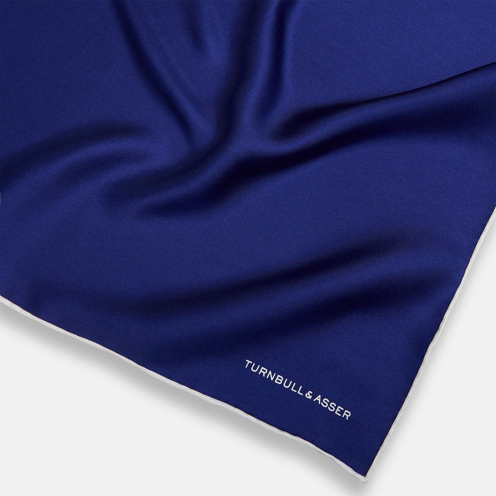 The Turnbull & Asser Royal Blue Piped Silk Pocket Square features white piping and the brand logo embroidered in white at the bottom right, making it perfect for formalwear.