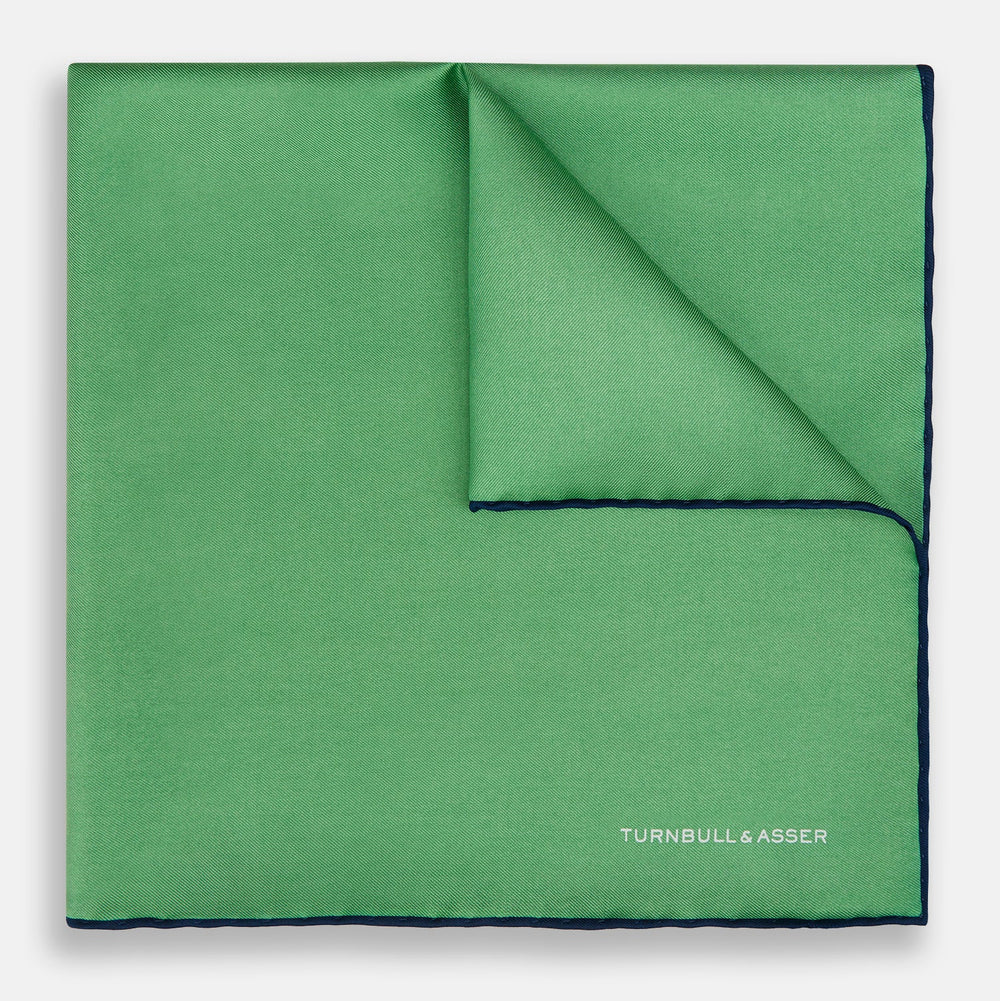 The Fern Green Piped Silk Pocket Square by Turnbull & Asser features a dark blue edge and white embroidered branding at the corner—an elegant accessory for formal wear.