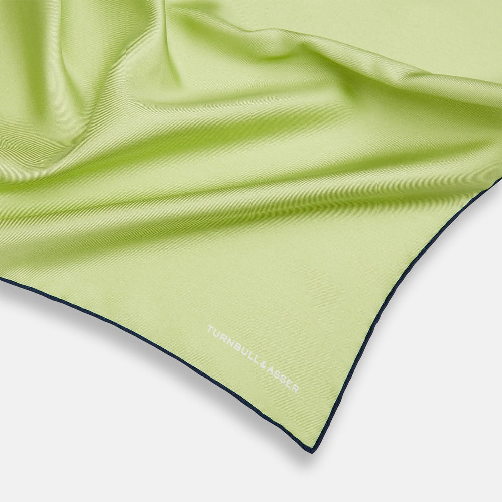 The Pistachio Green and Navy Piped Silk Pocket Square by Turnbull & Asser features crisp navy edging and an elegant brand logo, making it a refined addition to any formal outfit.