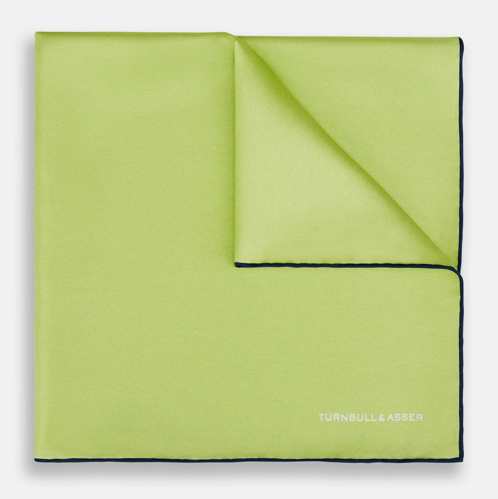 The Pistachio Green and Navy Piped Silk Pocket Square by Turnbull & Asser features crisp navy edging and an elegant brand logo, making it a refined addition to any formal outfit.