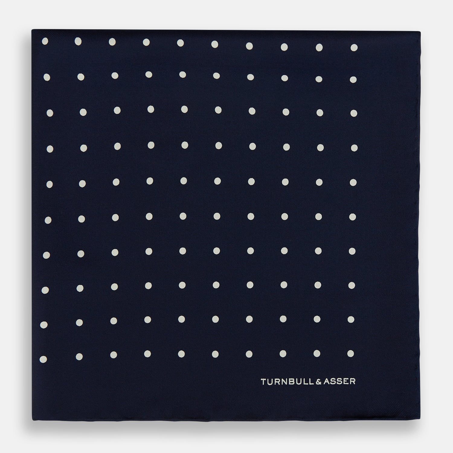 View 3 of product The Turnbull & Asser Navy and White Spot Silk Pocket Square features evenly spaced white polka dots and the brand logo in one corner—a stylish finishing touch for formal looks. One corner is neatly folded over.