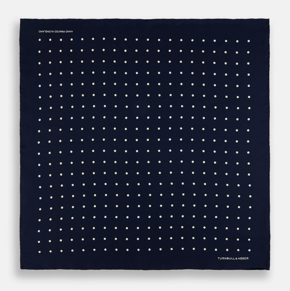 The Turnbull & Asser Navy and White Spot Silk Pocket Square features evenly spaced white polka dots and the brand logo in one corner—a stylish finishing touch for formal looks. One corner is neatly folded over.