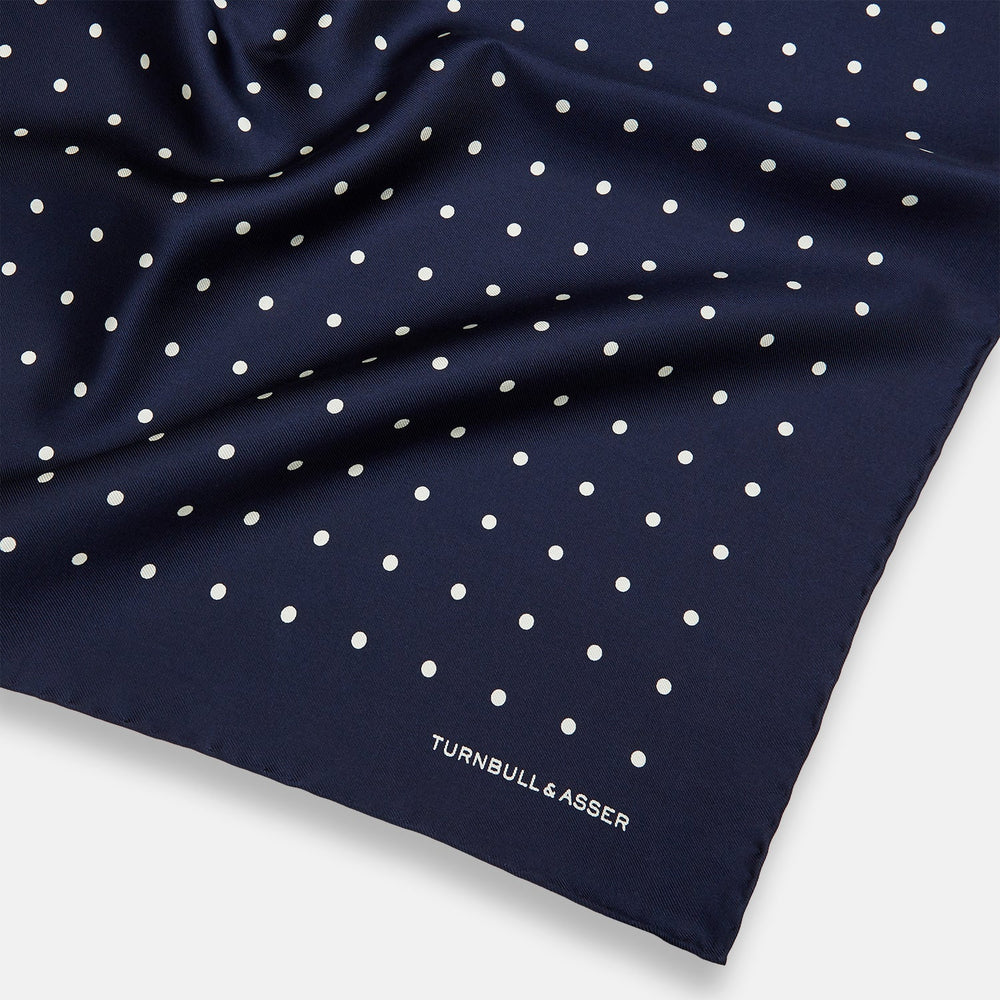 The Turnbull & Asser Navy and White Spot Silk Pocket Square features evenly spaced white polka dots and the brand logo in one corner—a stylish finishing touch for formal looks. One corner is neatly folded over.