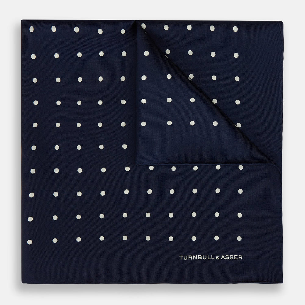 The Turnbull & Asser Navy and White Spot Silk Pocket Square features evenly spaced white polka dots and the brand logo in one corner—a stylish finishing touch for formal looks. One corner is neatly folded over.