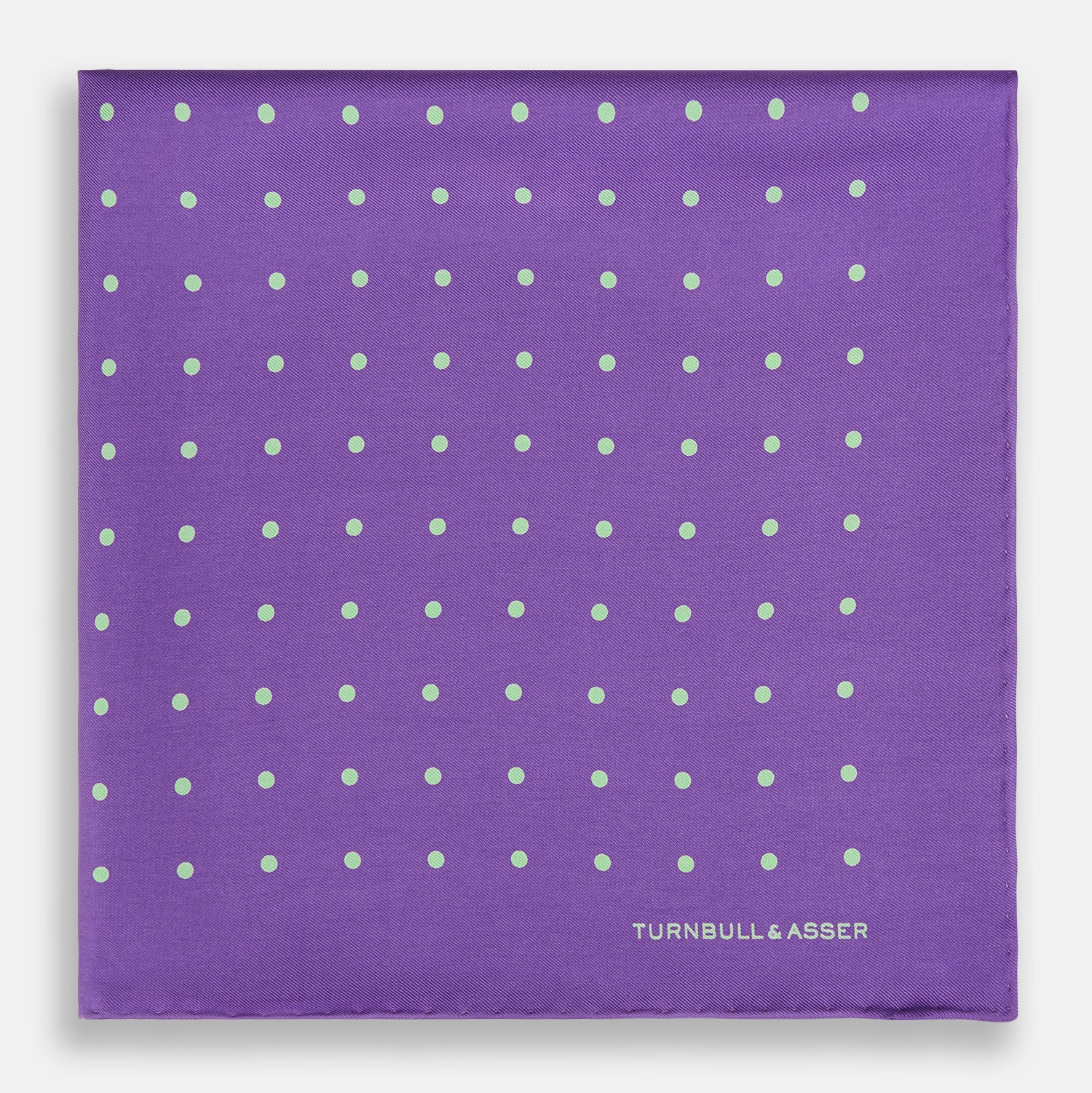 View 3 of product The PURPLE AND MINT SPOT SILK POCKET SQUARE by Turnbull & Asser features mint spots on purple silk and the brand's white logo in the lower right corner. One corner is folded over, perfect for elevating any gentleman’s formal look.