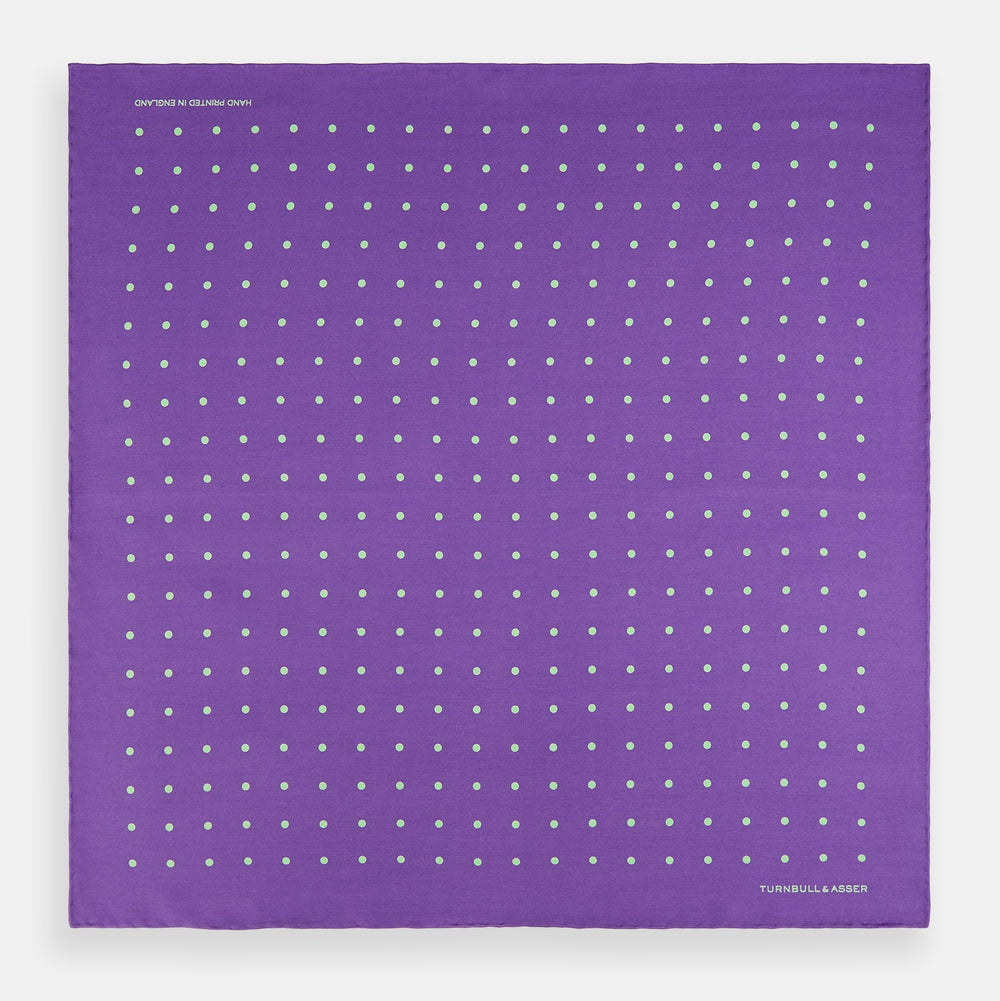 The PURPLE AND MINT SPOT SILK POCKET SQUARE by Turnbull & Asser features mint spots on purple silk and the brand's white logo in the lower right corner. One corner is folded over, perfect for elevating any gentleman’s formal look.