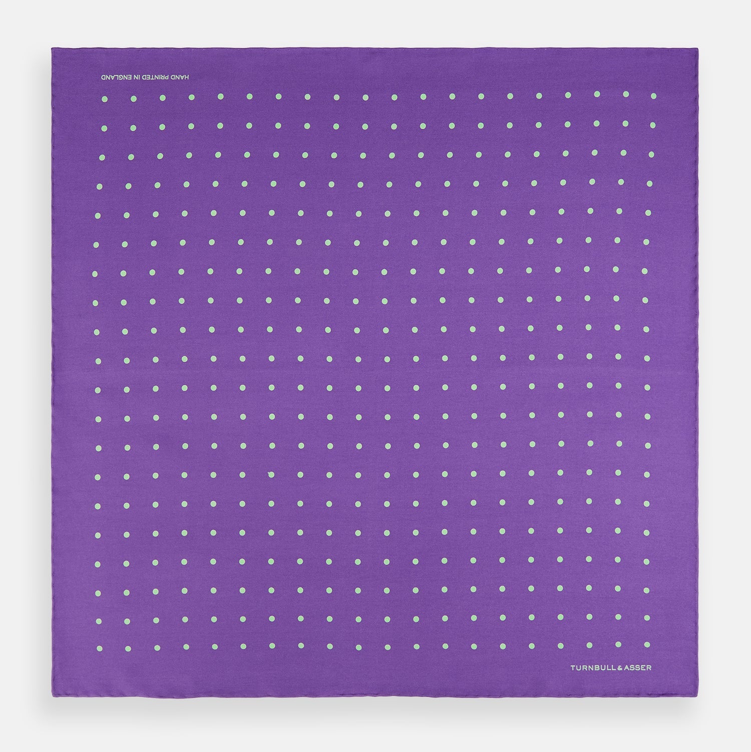 View 4 of product The PURPLE AND MINT SPOT SILK POCKET SQUARE by Turnbull & Asser features mint spots on purple silk and the brand's white logo in the lower right corner. One corner is folded over, perfect for elevating any gentleman’s formal look.