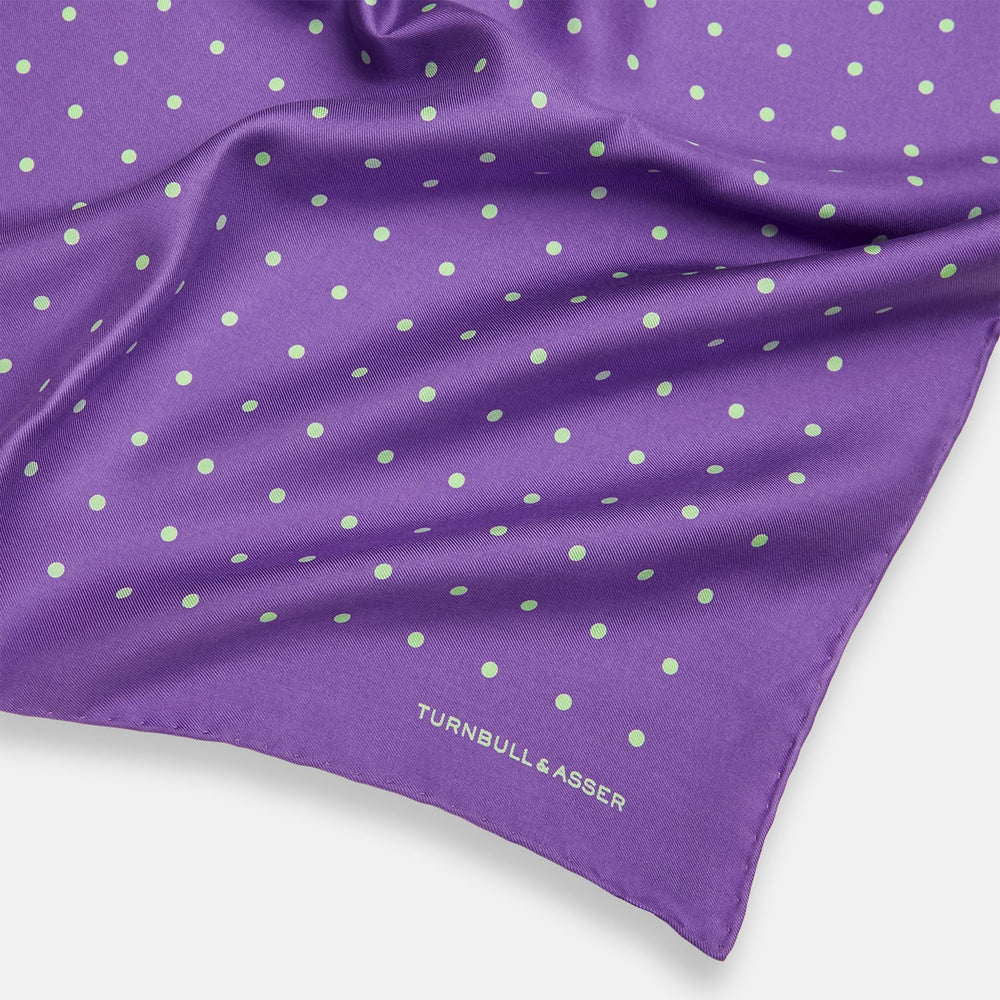 The PURPLE AND MINT SPOT SILK POCKET SQUARE by Turnbull & Asser features mint spots on purple silk and the brand's white logo in the lower right corner. One corner is folded over, perfect for elevating any gentleman’s formal look.