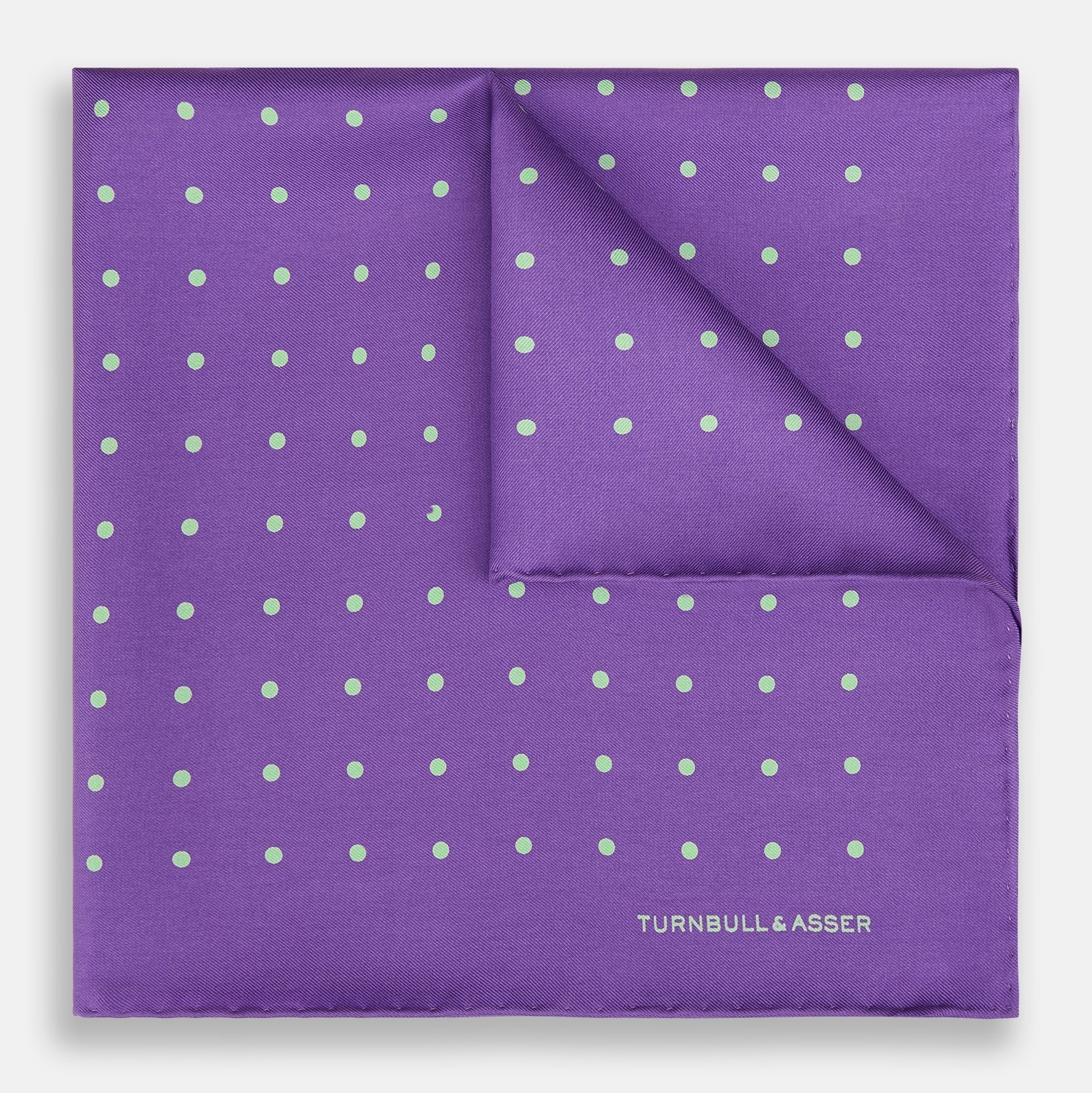 View 1 of product The PURPLE AND MINT SPOT SILK POCKET SQUARE by Turnbull & Asser features mint spots on purple silk and the brand's white logo in the lower right corner. One corner is folded over, perfect for elevating any gentleman’s formal look.