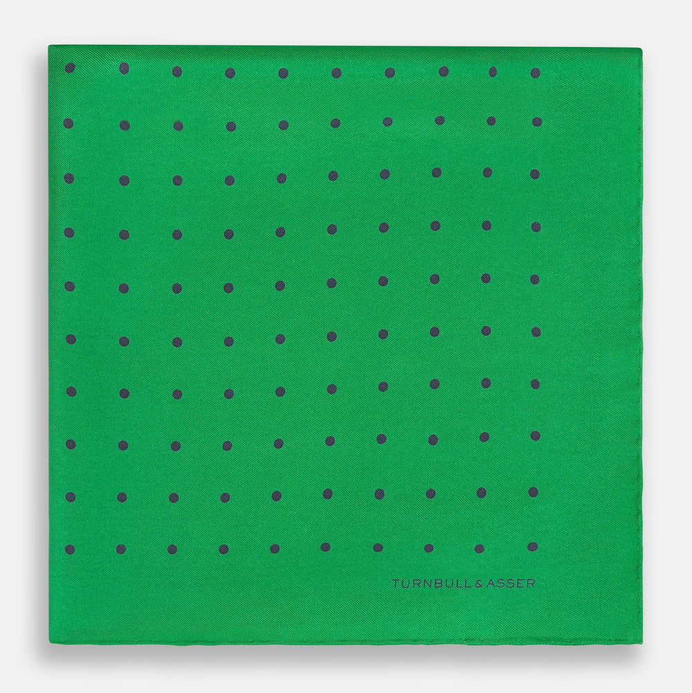 The Green and Navy Spot Silk Pocket Square by Turnbull & Asser features evenly spaced navy spots on green silk, with the brand name printed in the lower right corner and one corner folded over. Ideal for formal wear.