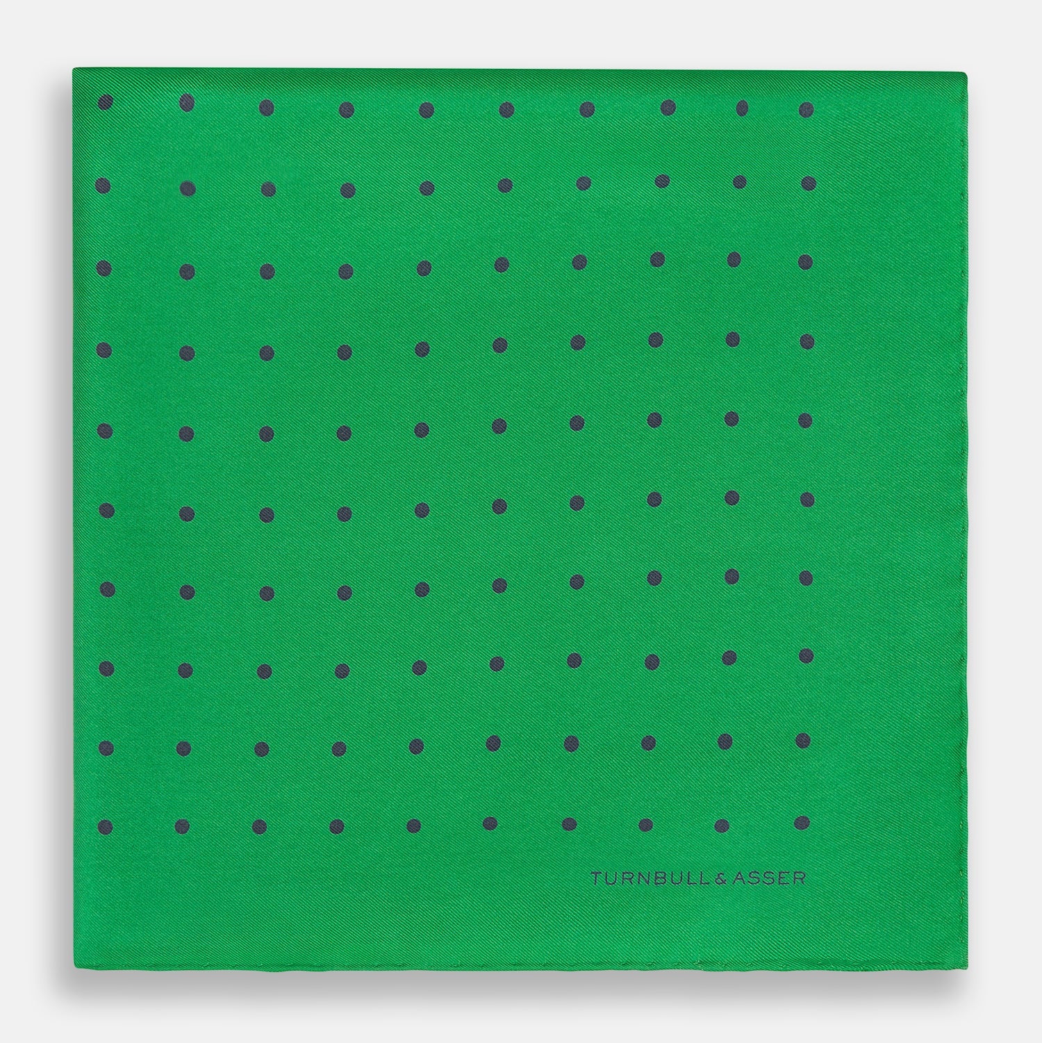 View 3 of product The Green and Navy Spot Silk Pocket Square by Turnbull & Asser features evenly spaced navy spots on green silk, with the brand name printed in the lower right corner and one corner folded over. Ideal for formal wear.