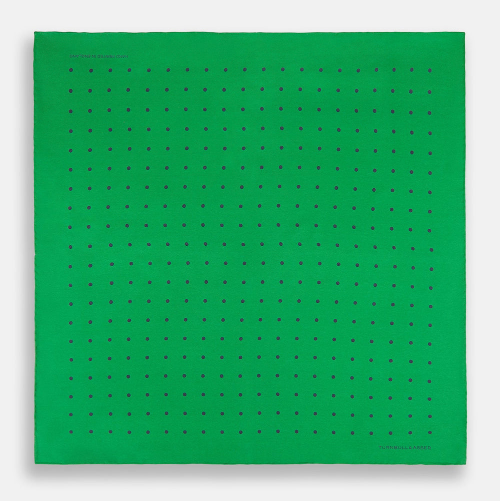 The Green and Navy Spot Silk Pocket Square by Turnbull & Asser features evenly spaced navy spots on green silk, with the brand name printed in the lower right corner and one corner folded over. Ideal for formal wear.