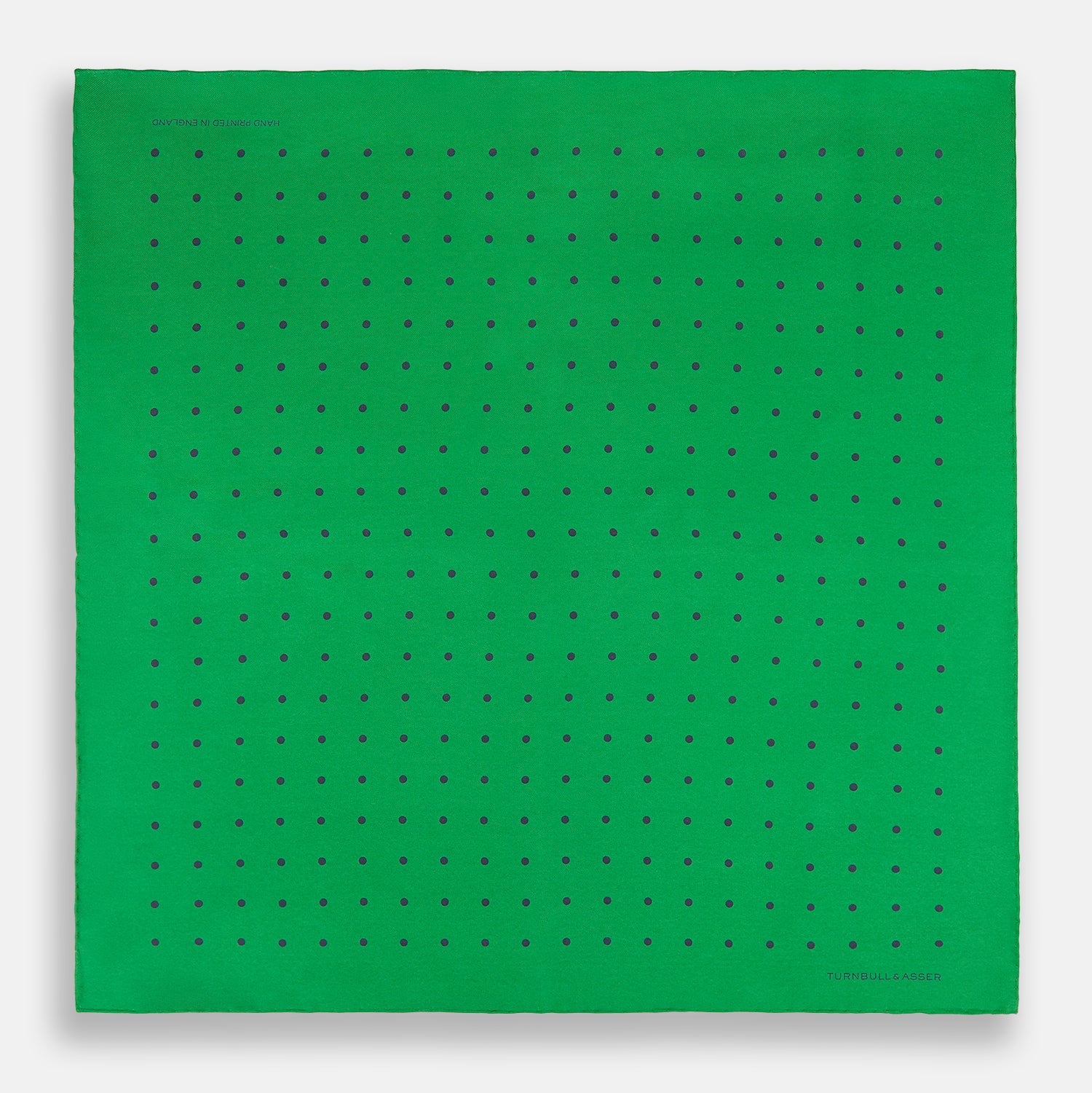 View 4 of product The Green and Navy Spot Silk Pocket Square by Turnbull & Asser features evenly spaced navy spots on green silk, with the brand name printed in the lower right corner and one corner folded over. Ideal for formal wear.