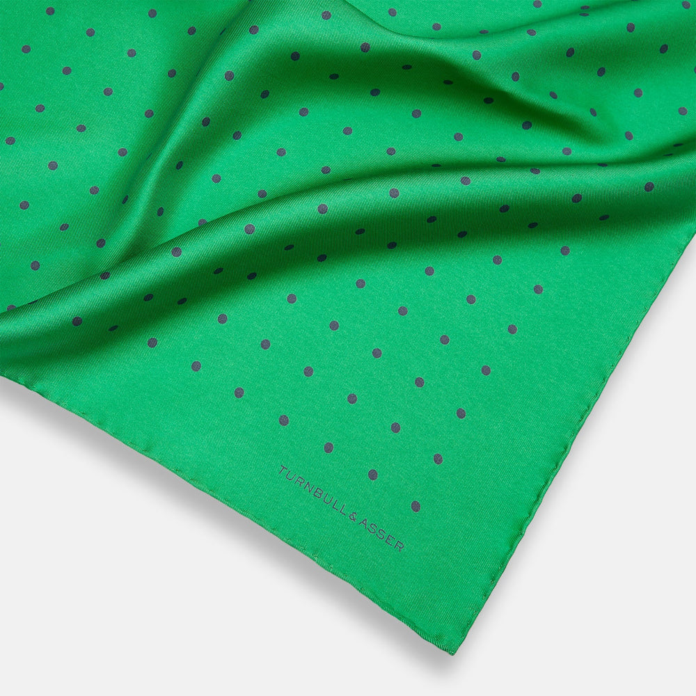 The Green and Navy Spot Silk Pocket Square by Turnbull & Asser features evenly spaced navy spots on green silk, with the brand name printed in the lower right corner and one corner folded over. Ideal for formal wear.