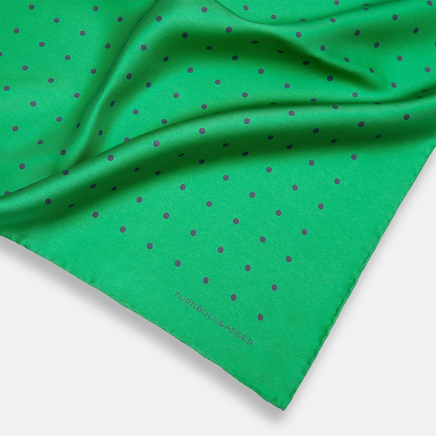 View 2 of product The Green and Navy Spot Silk Pocket Square by Turnbull & Asser features evenly spaced navy spots on green silk, with the brand name printed in the lower right corner and one corner folded over. Ideal for formal wear.