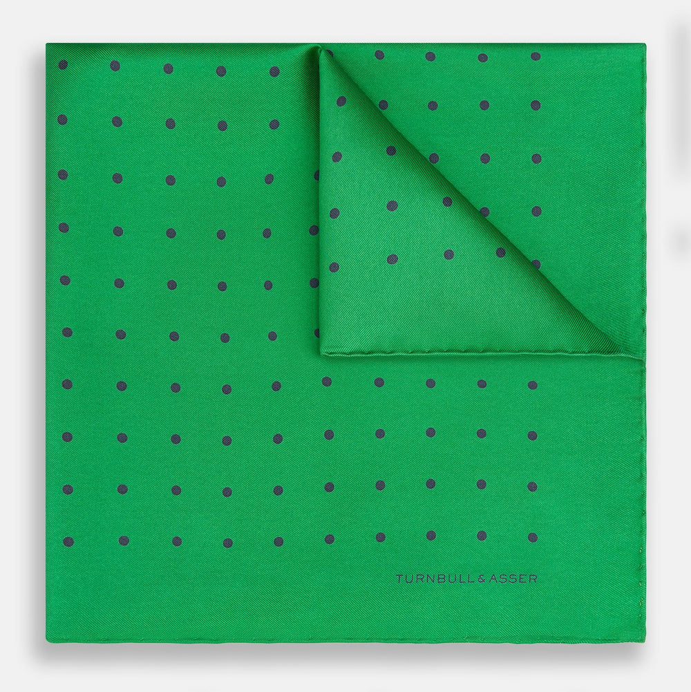 The Green and Navy Spot Silk Pocket Square by Turnbull & Asser features evenly spaced navy spots on green silk, with the brand name printed in the lower right corner and one corner folded over. Ideal for formal wear.