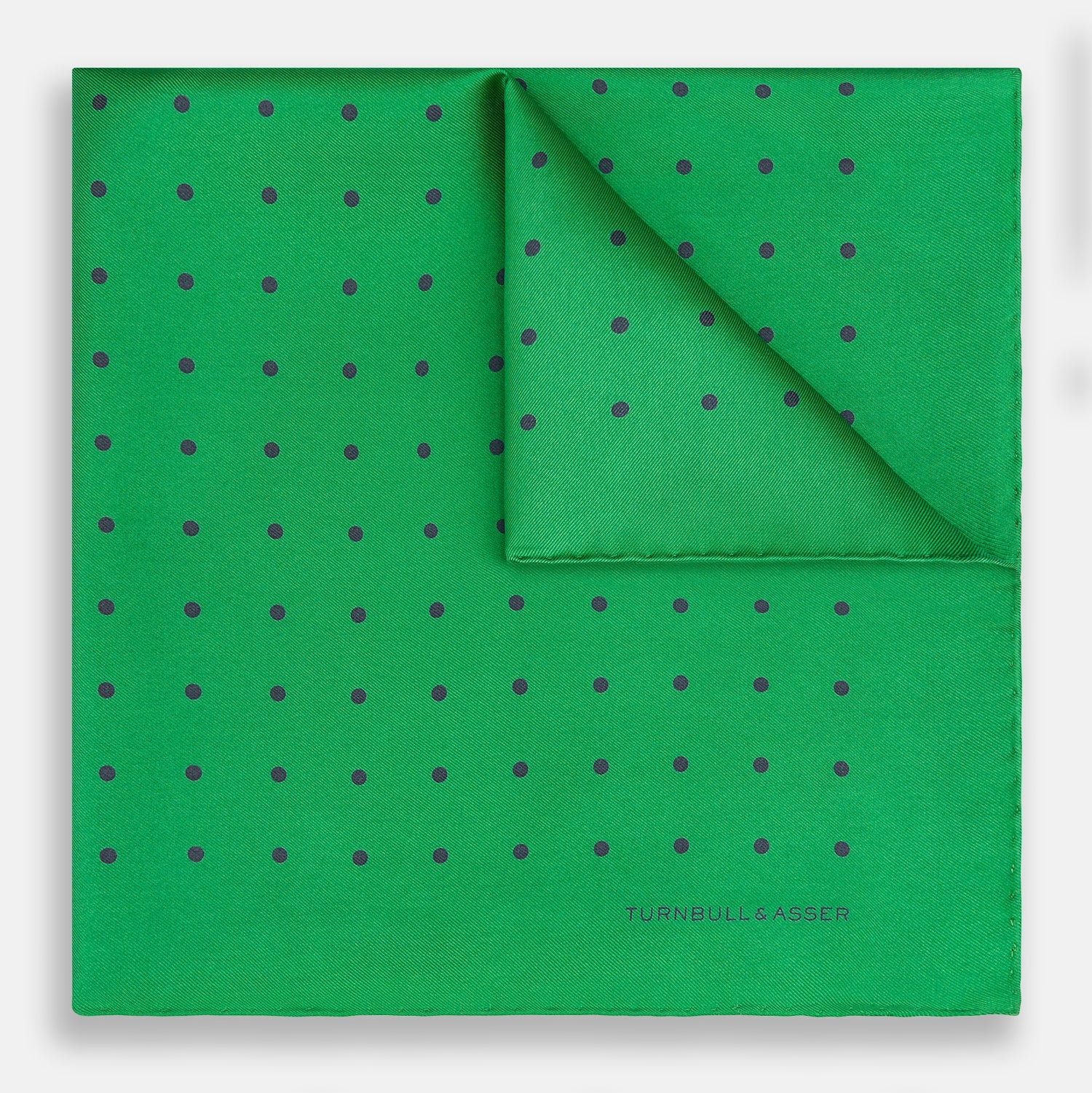 View 1 of product The Green and Navy Spot Silk Pocket Square by Turnbull & Asser features evenly spaced navy spots on green silk, with the brand name printed in the lower right corner and one corner folded over. Ideal for formal wear.
