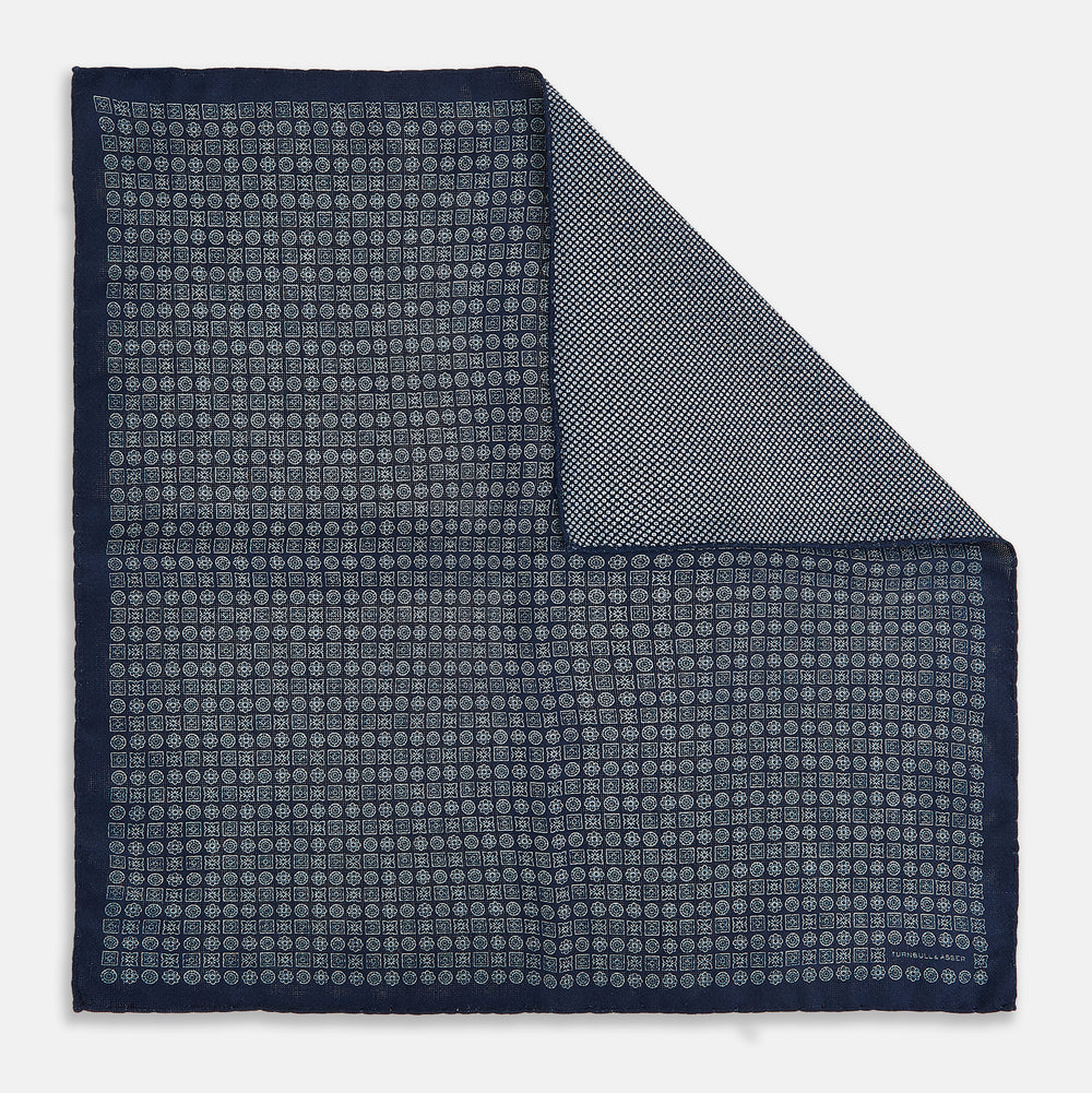 The Rosette Navy Pocket Square in Silk by Turnbull & Asser UK is a square, navy blue accessory featuring a small geometric pattern. Shown laid flat with one corner folded, it reveals a contrasting, finer motif on the reverse side. This elegant double-sided design offers versatile styling options for any occasion.