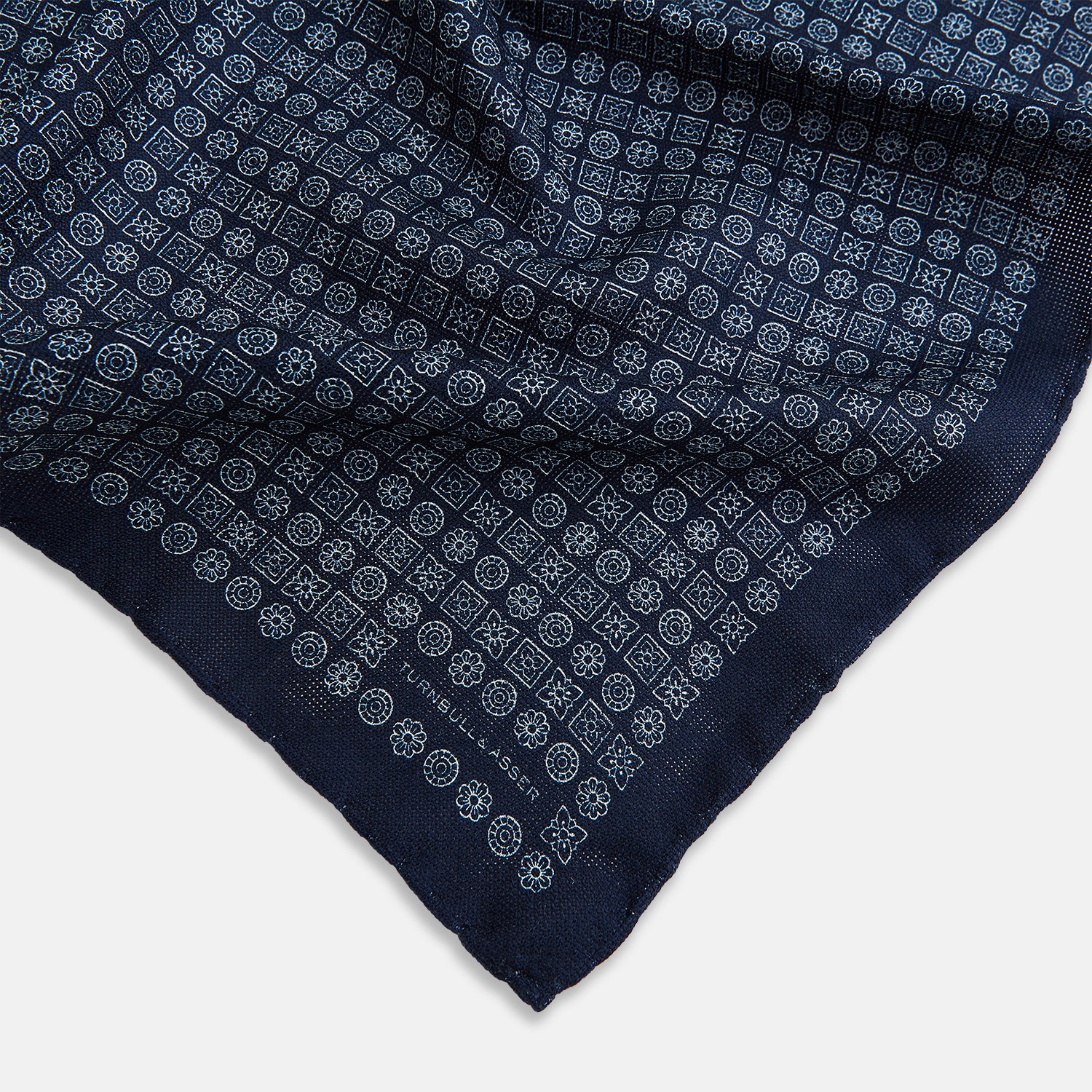 View 2 of product The Rosette Navy Pocket Square in Silk by Turnbull & Asser UK is a square, navy blue accessory featuring a small geometric pattern. Shown laid flat with one corner folded, it reveals a contrasting, finer motif on the reverse side. This elegant double-sided design offers versatile styling options for any occasion.