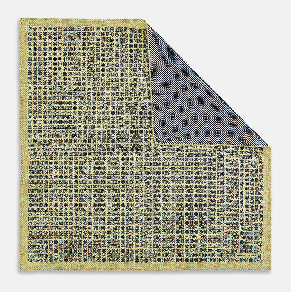 The Rosette Yellow Pocket Square in Silk by Turnbull & Asser UK features a square design with a yellow border, adorned with a grid of small black and white circles. One corner is neatly folded to reveal a reverse side in a refined black and white micro-checkered pattern, while subtle geometric medallions lend an added touch of elegance to the overall look.
