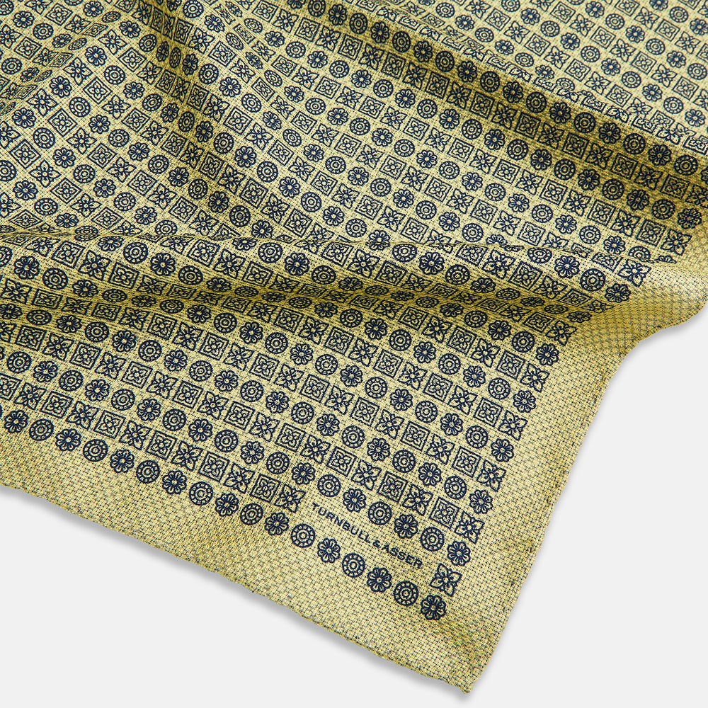 The Rosette Yellow Pocket Square in Silk by Turnbull & Asser UK features a square design with a yellow border, adorned with a grid of small black and white circles. One corner is neatly folded to reveal a reverse side in a refined black and white micro-checkered pattern, while subtle geometric medallions lend an added touch of elegance to the overall look.