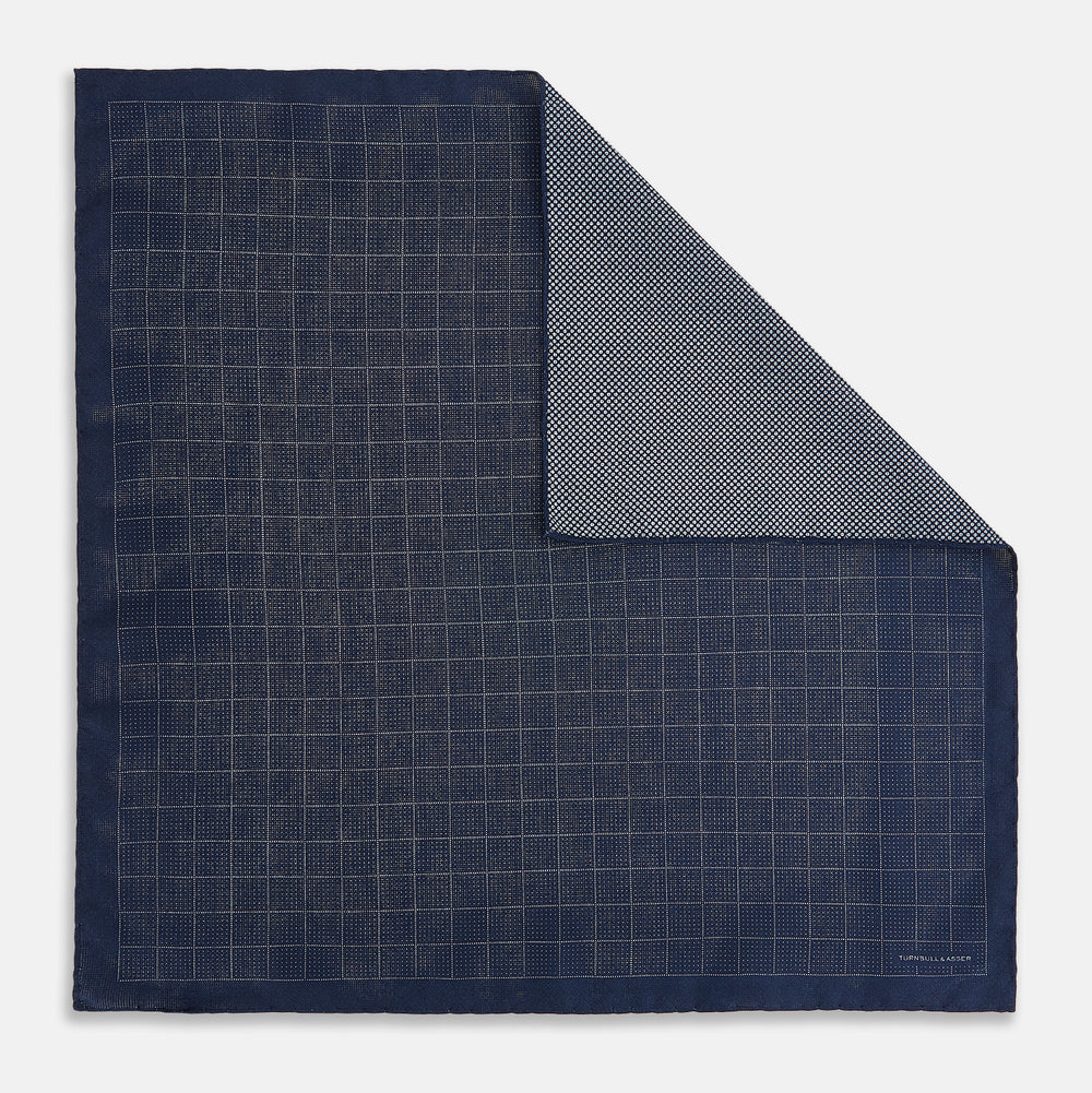 The Checked Blue Pocket Square in Silk by Turnbull & Asser UK features a navy base with a light blue grid pattern on one side and a refined dotted design on the other. This double-sided accessory is shown partially folded to elegantly showcase both distinctive patterns.