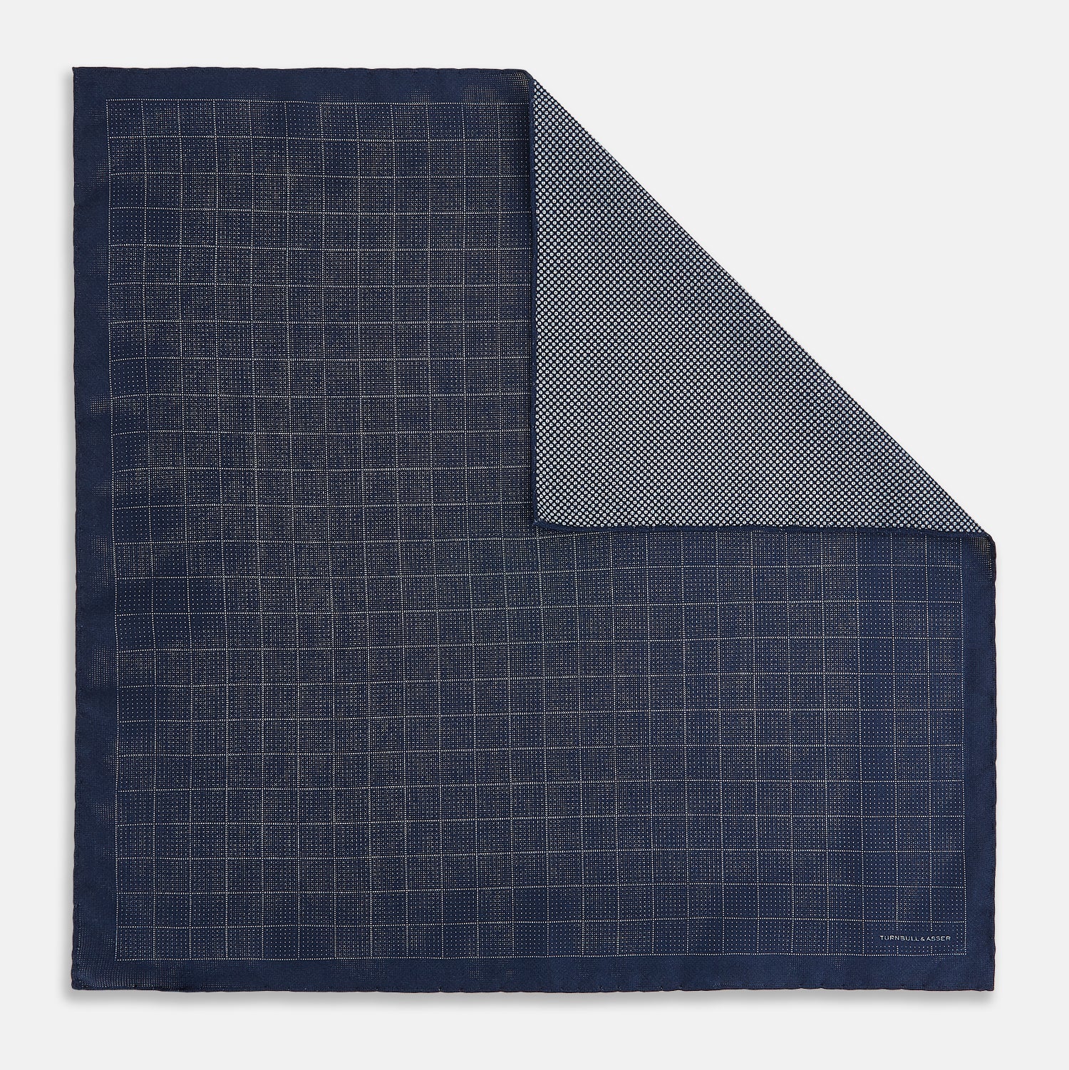 View 1 of product The Checked Blue Pocket Square in Silk by Turnbull & Asser UK features a navy base with a light blue grid pattern on one side and a refined dotted design on the other. This double-sided accessory is shown partially folded to elegantly showcase both distinctive patterns.