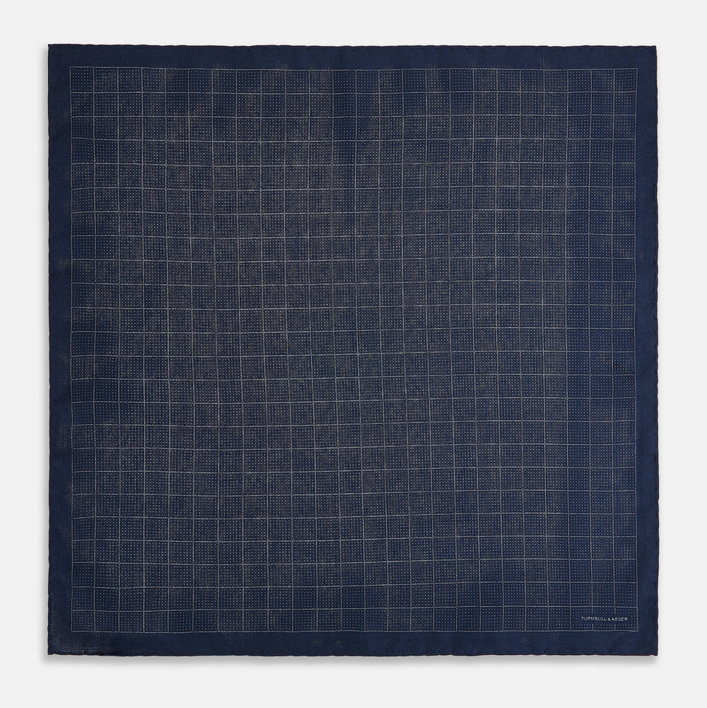 The Checked Blue Pocket Square in Silk by Turnbull & Asser UK features a navy base with a light blue grid pattern on one side and a refined dotted design on the other. This double-sided accessory is shown partially folded to elegantly showcase both distinctive patterns.