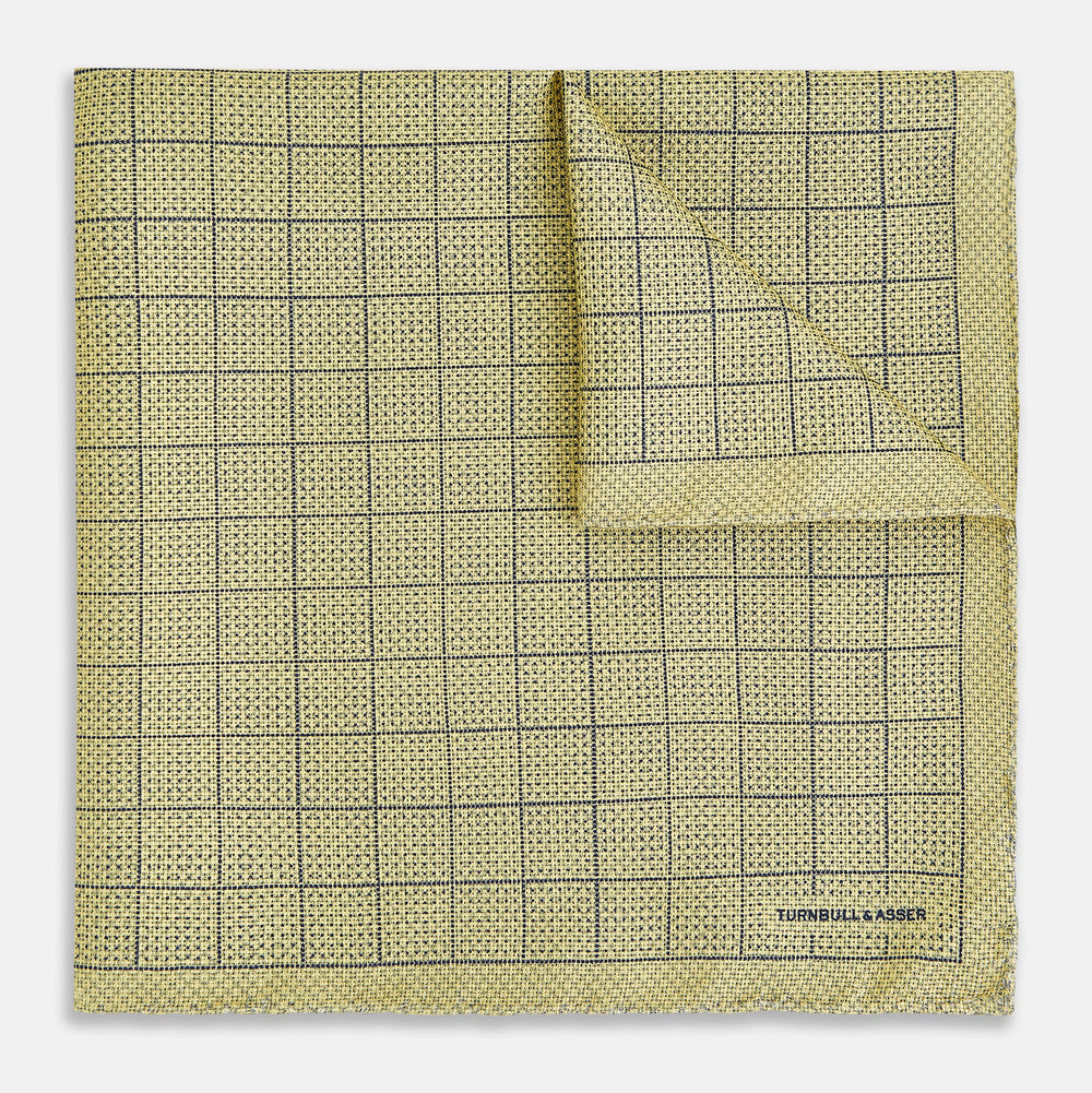 A yellow silk pocket square featuring a small black grid pattern of dotted squares, neatly folded at one corner. "Turnbull & Asser UK" is printed in the bottom right corner.