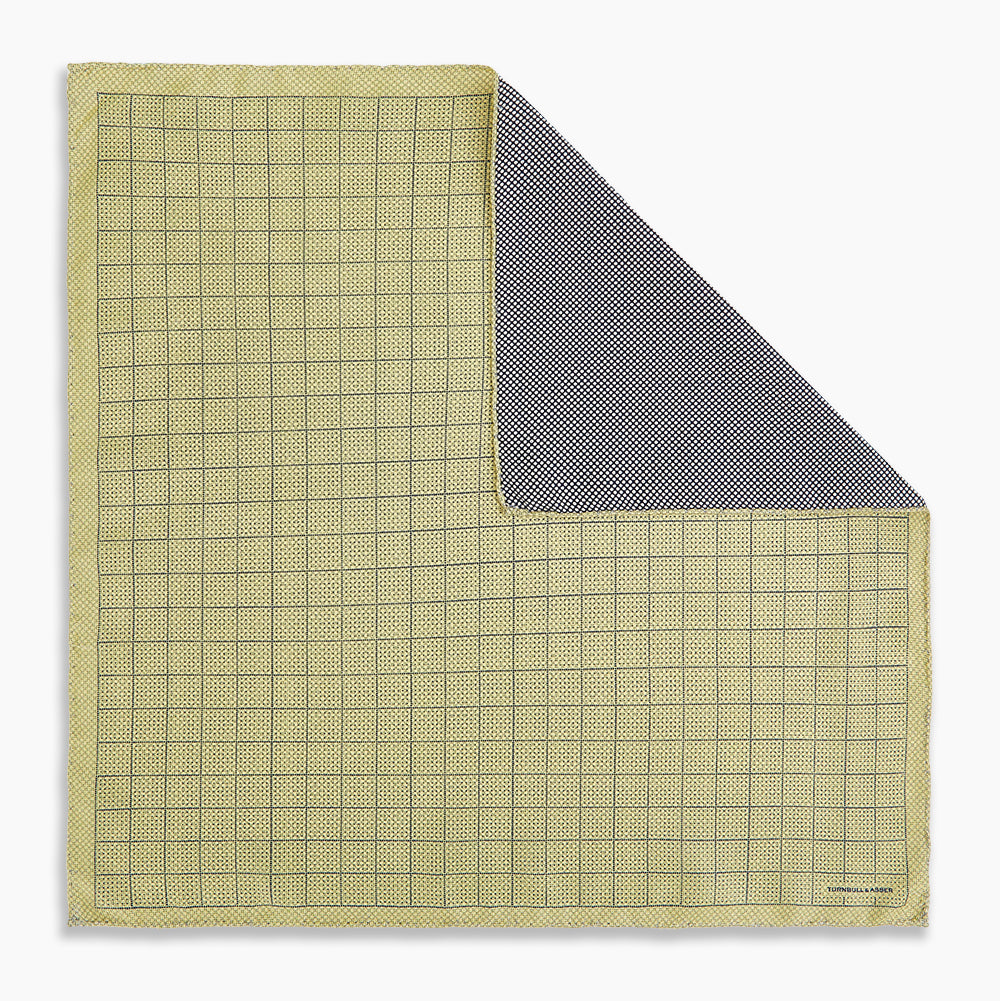 A yellow silk pocket square featuring a small black grid pattern of dotted squares, neatly folded at one corner. "Turnbull & Asser UK" is printed in the bottom right corner.