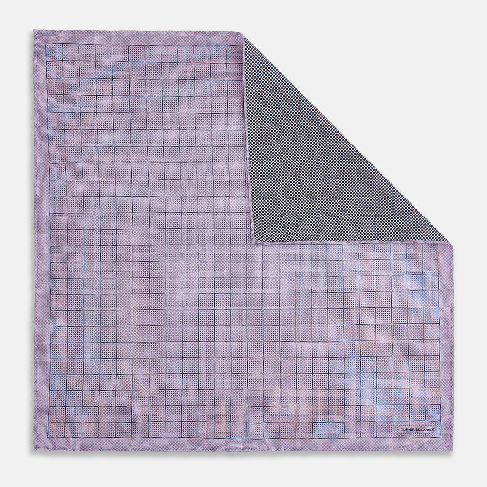 The Checked Lilac Pocket Square in Silk by Turnbull & Asser UK features a grid pattern in purple with a solid border, neatly folded to display its double-sided design and contrasting black and white dotted squares on the reverse side.