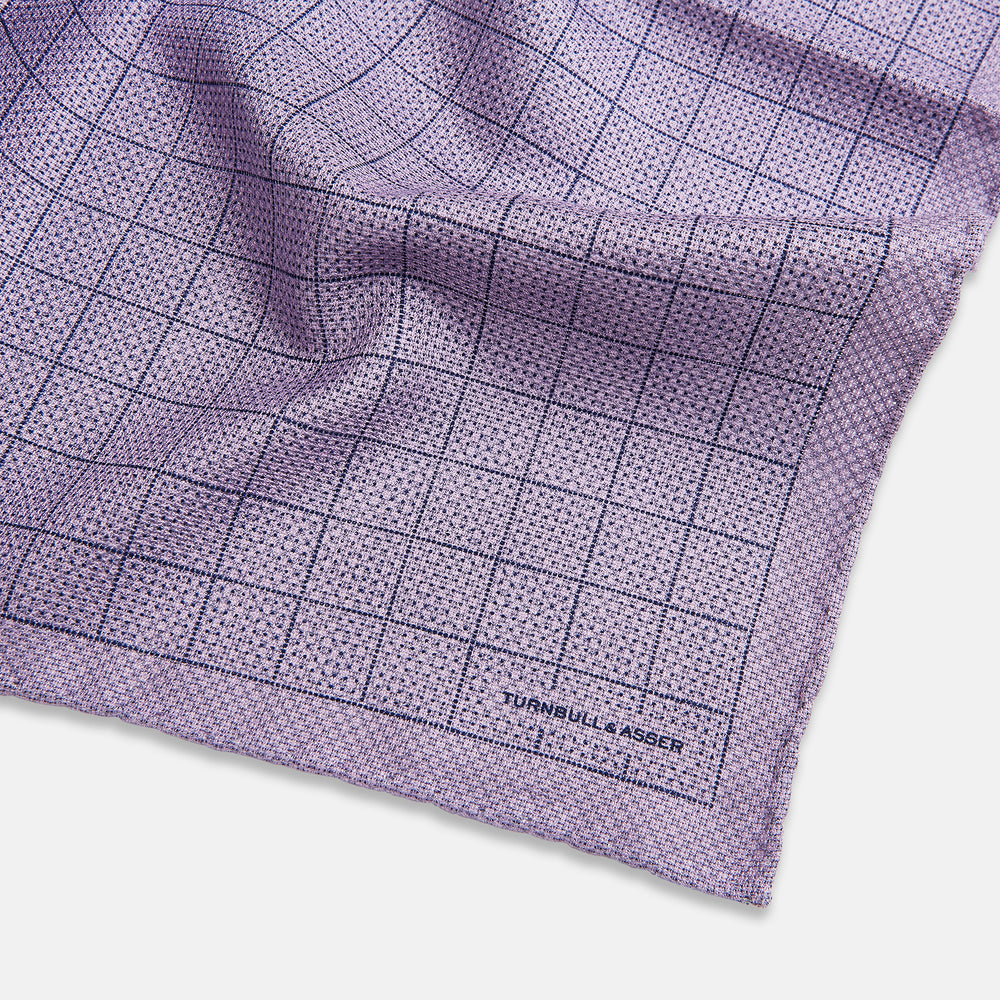 The Checked Lilac Pocket Square in Silk by Turnbull & Asser UK features a grid pattern in purple with a solid border, neatly folded to display its double-sided design and contrasting black and white dotted squares on the reverse side.