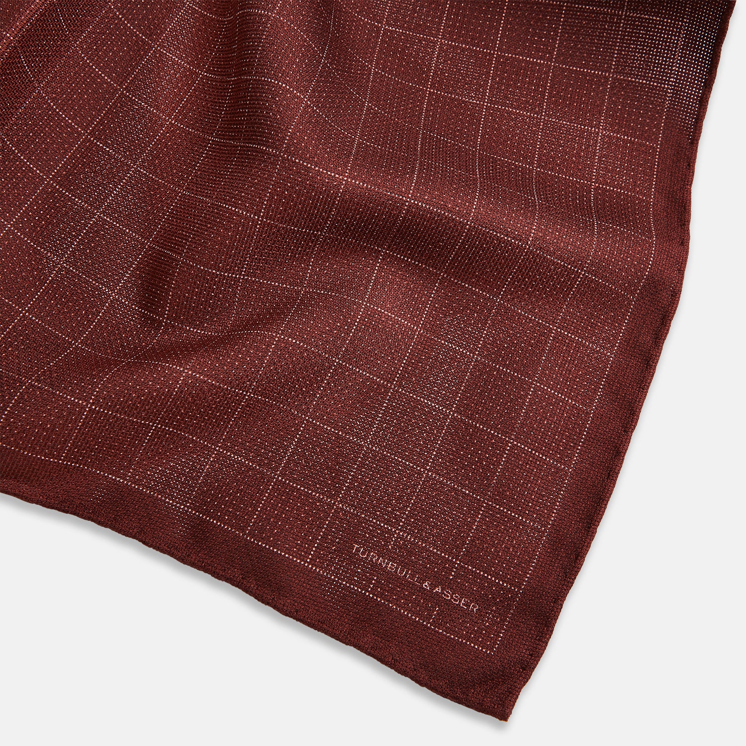 View 3 of product The Turnbull & Asser UK Checked Burgundy Pocket Square in Silk features a sophisticated burgundy hue with a subtle grid pattern. It's displayed folded at one corner to reveal a contrasting small checkered design on the reverse, showcasing its refined double-sided craftsmanship.