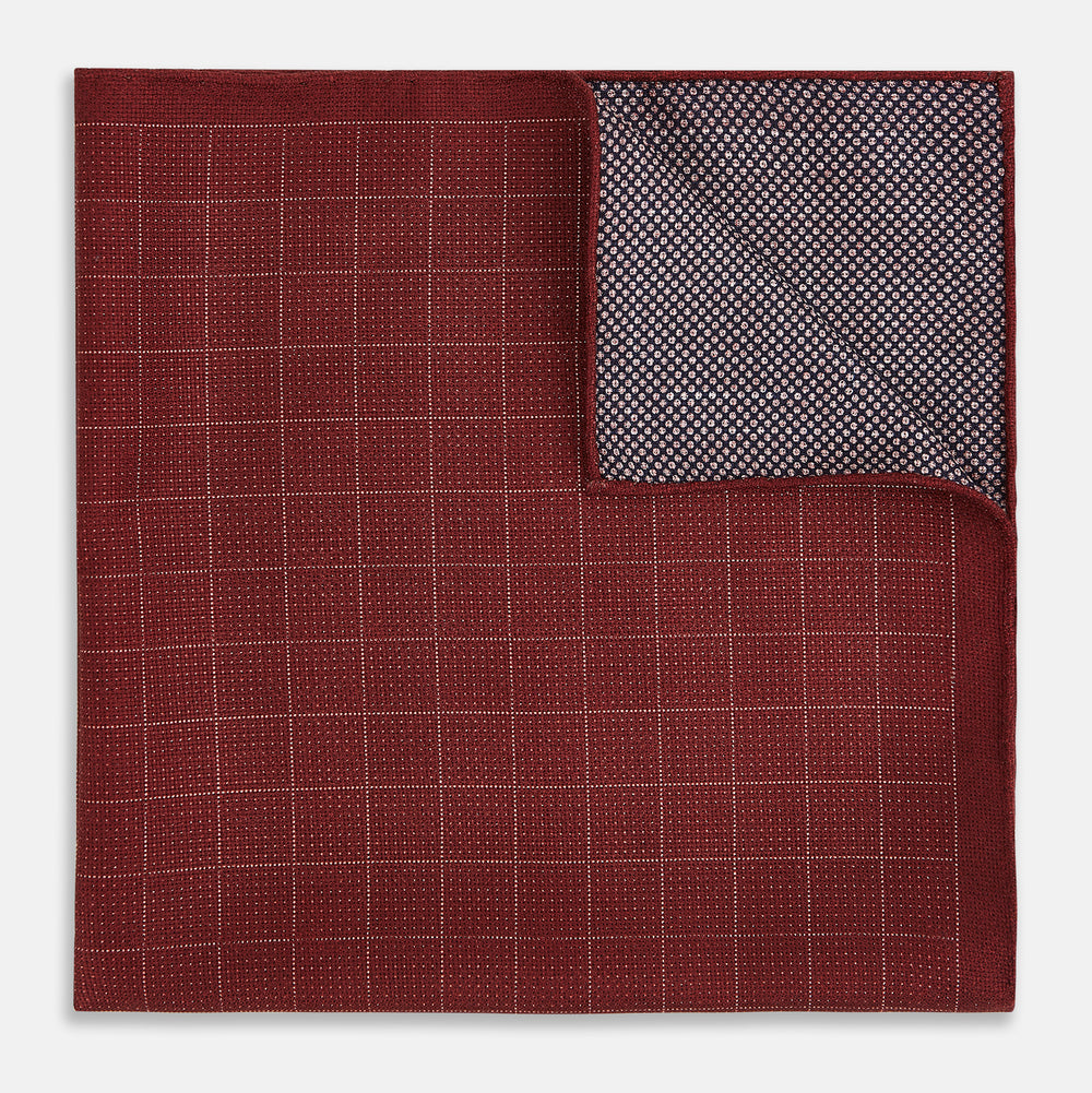 The Turnbull & Asser UK Checked Burgundy Pocket Square in Silk features a sophisticated burgundy hue with a subtle grid pattern. It's displayed folded at one corner to reveal a contrasting small checkered design on the reverse, showcasing its refined double-sided craftsmanship.