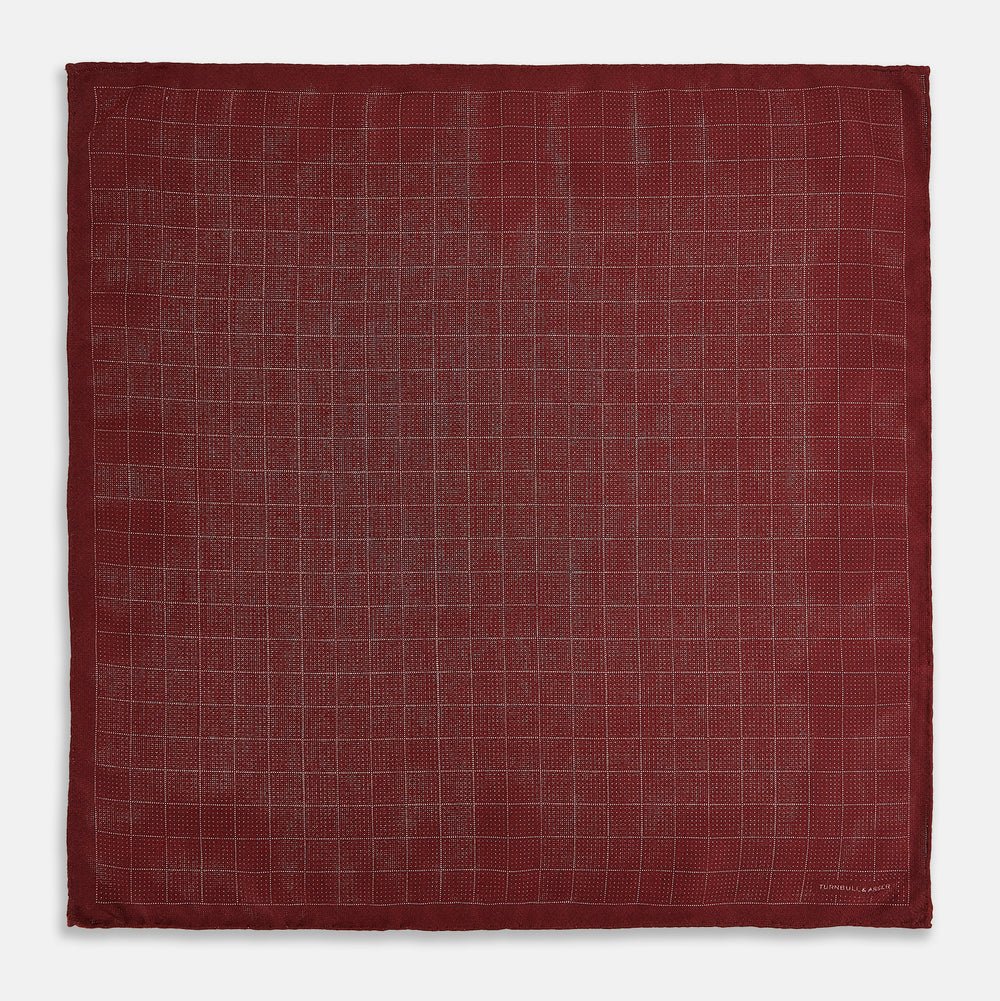 The Turnbull & Asser UK Checked Burgundy Pocket Square in Silk features a sophisticated burgundy hue with a subtle grid pattern. It's displayed folded at one corner to reveal a contrasting small checkered design on the reverse, showcasing its refined double-sided craftsmanship.