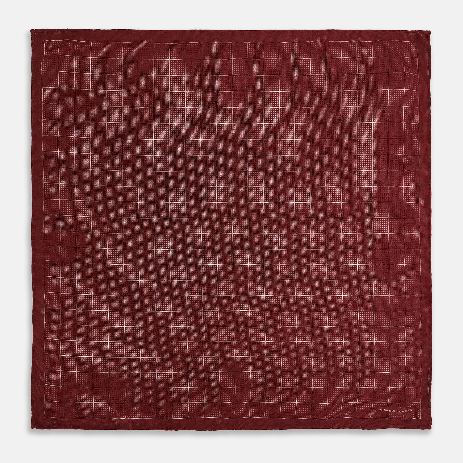 View 5 of product The Turnbull & Asser UK Checked Burgundy Pocket Square in Silk features a sophisticated burgundy hue with a subtle grid pattern. It's displayed folded at one corner to reveal a contrasting small checkered design on the reverse, showcasing its refined double-sided craftsmanship.