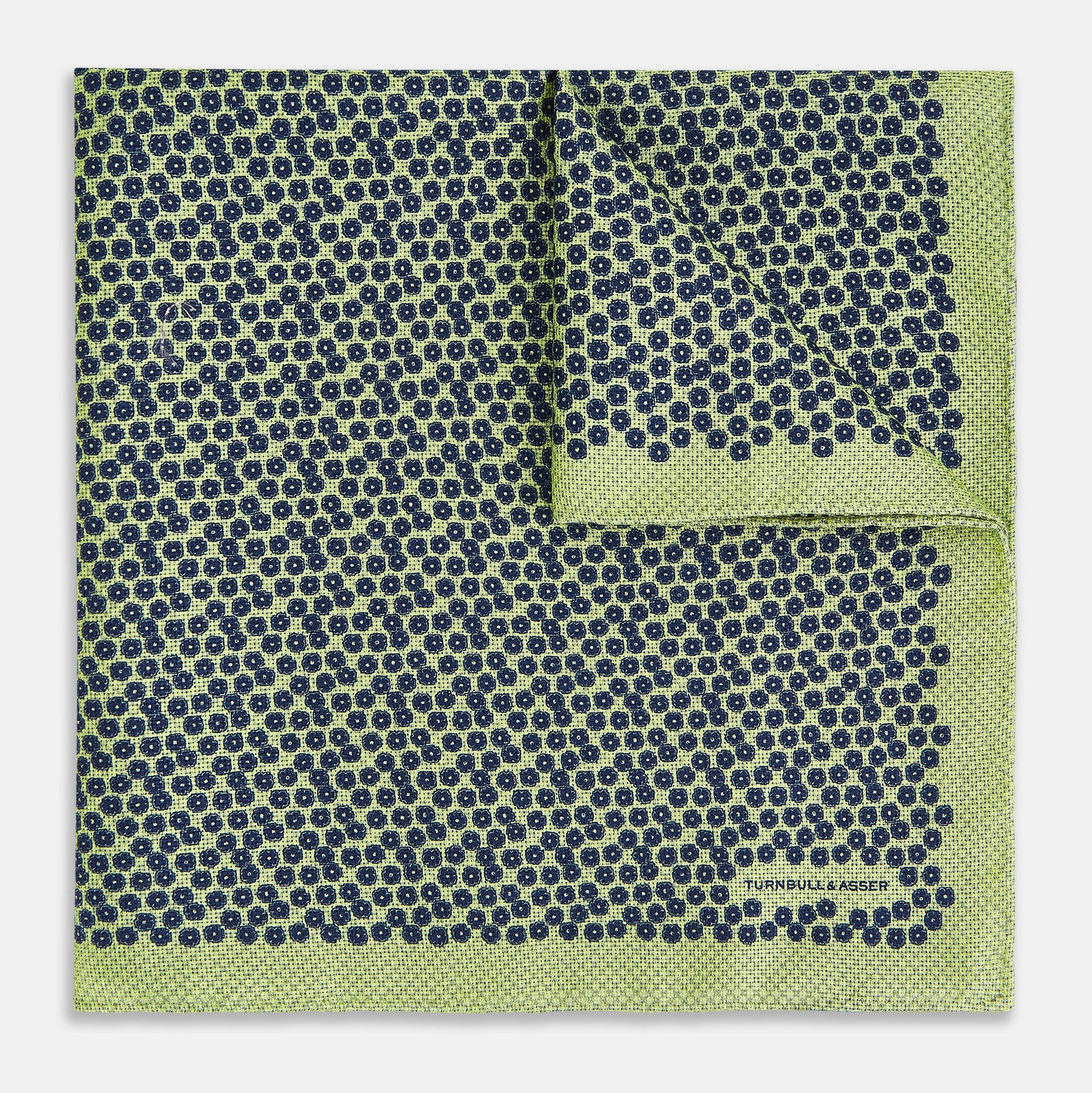 View 2 of product The Turnbull & Asser UK Floral Green Pocket Square in Silk is a square accessory featuring a blue floral pattern with a green border, partly folded at one corner to showcase its double-sided design, revealing a navy and light green dotted pattern on the reverse.