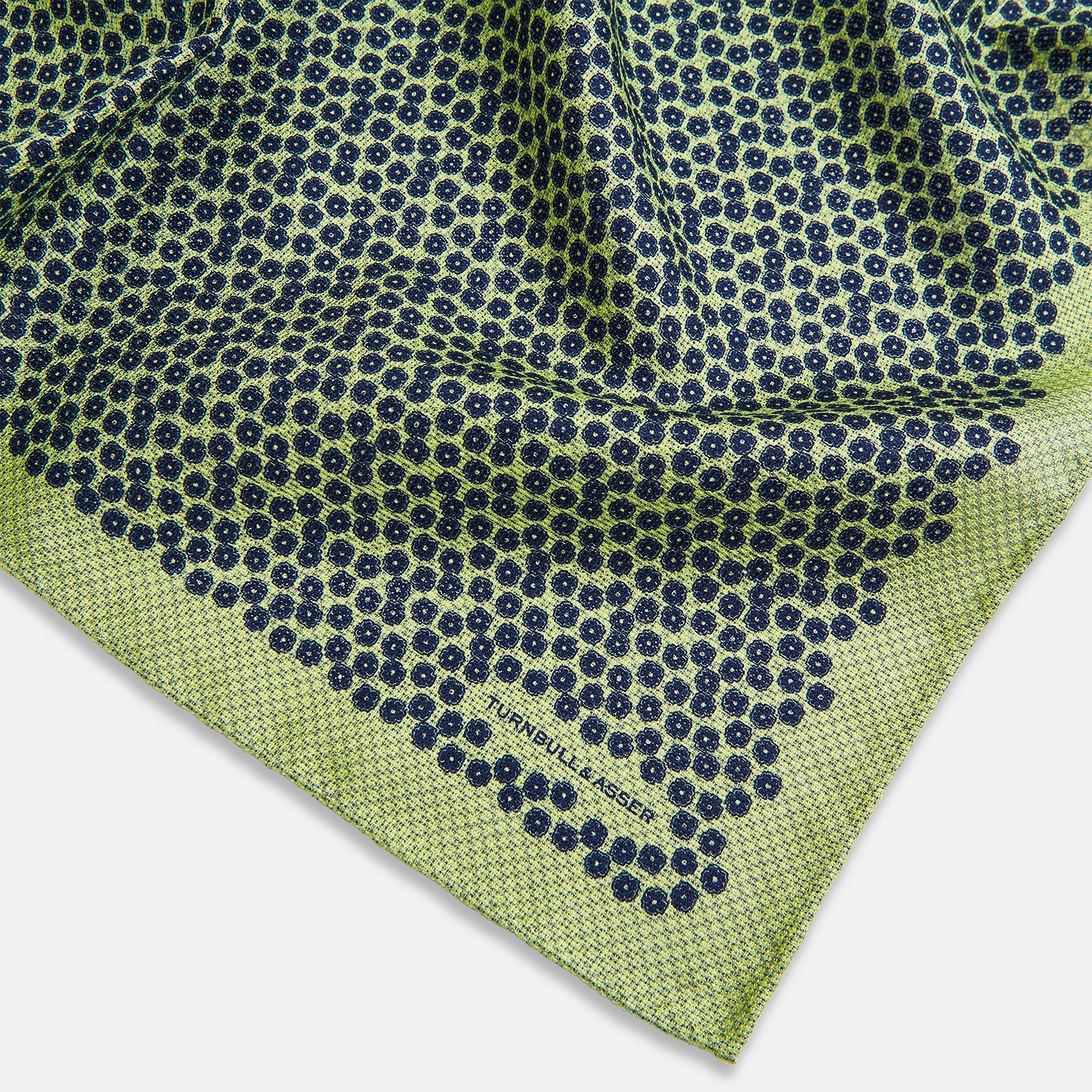 View 3 of product The Turnbull & Asser UK Floral Green Pocket Square in Silk is a square accessory featuring a blue floral pattern with a green border, partly folded at one corner to showcase its double-sided design, revealing a navy and light green dotted pattern on the reverse.