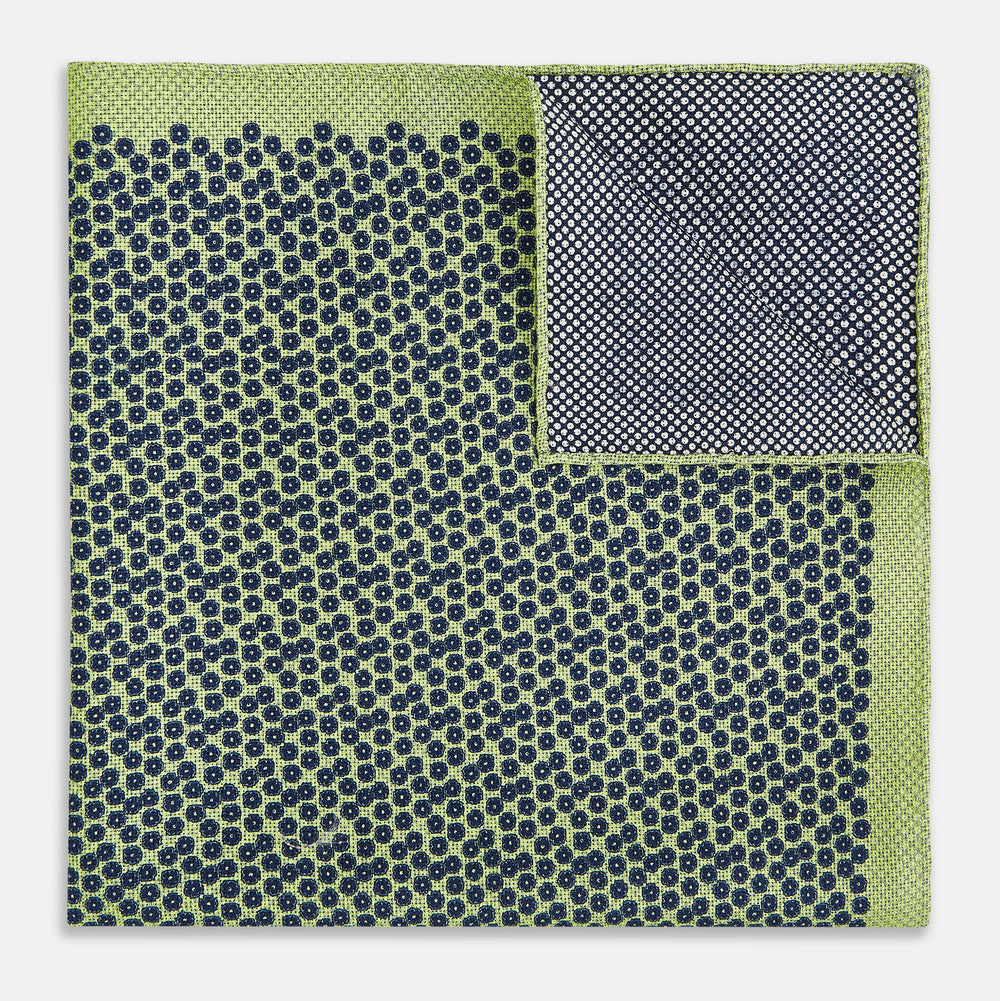 The Turnbull & Asser UK Floral Green Pocket Square in Silk is a square accessory featuring a blue floral pattern with a green border, partly folded at one corner to showcase its double-sided design, revealing a navy and light green dotted pattern on the reverse.