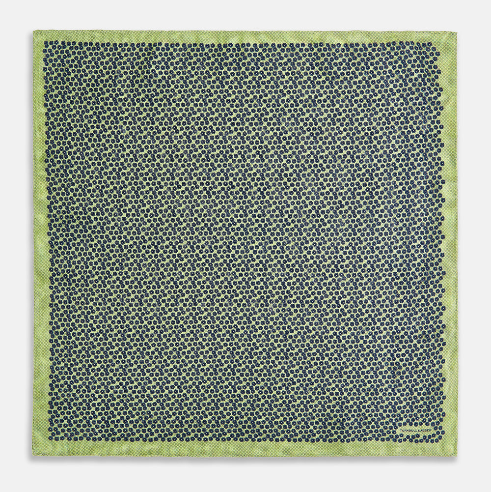 The Turnbull & Asser UK Floral Green Pocket Square in Silk is a square accessory featuring a blue floral pattern with a green border, partly folded at one corner to showcase its double-sided design, revealing a navy and light green dotted pattern on the reverse.