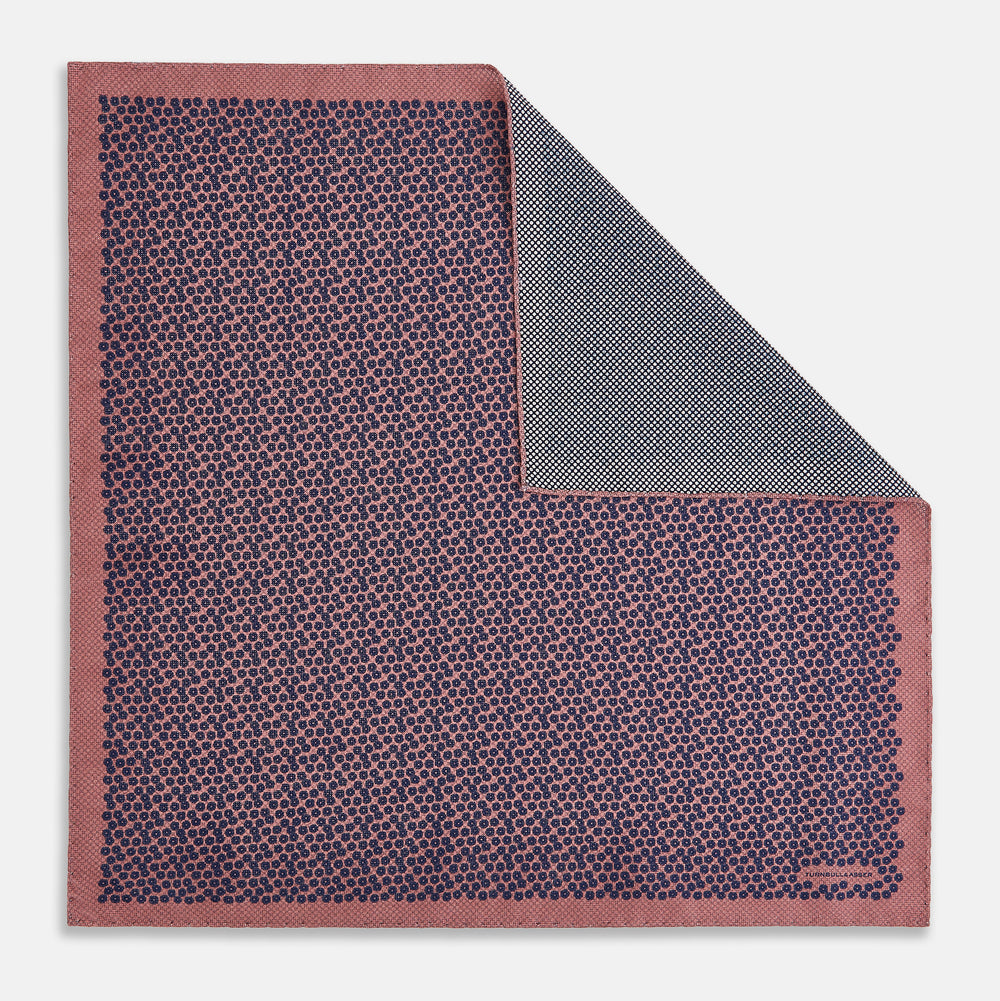 The Floral Pink Pocket Square in Silk by Turnbull & Asser UK showcases a refined pattern with a pink floral design and a contrasting border. The top right corner is elegantly folded to reveal the reverse side’s complementary blue and white dotted motif, emphasizing its double-sided versatility.