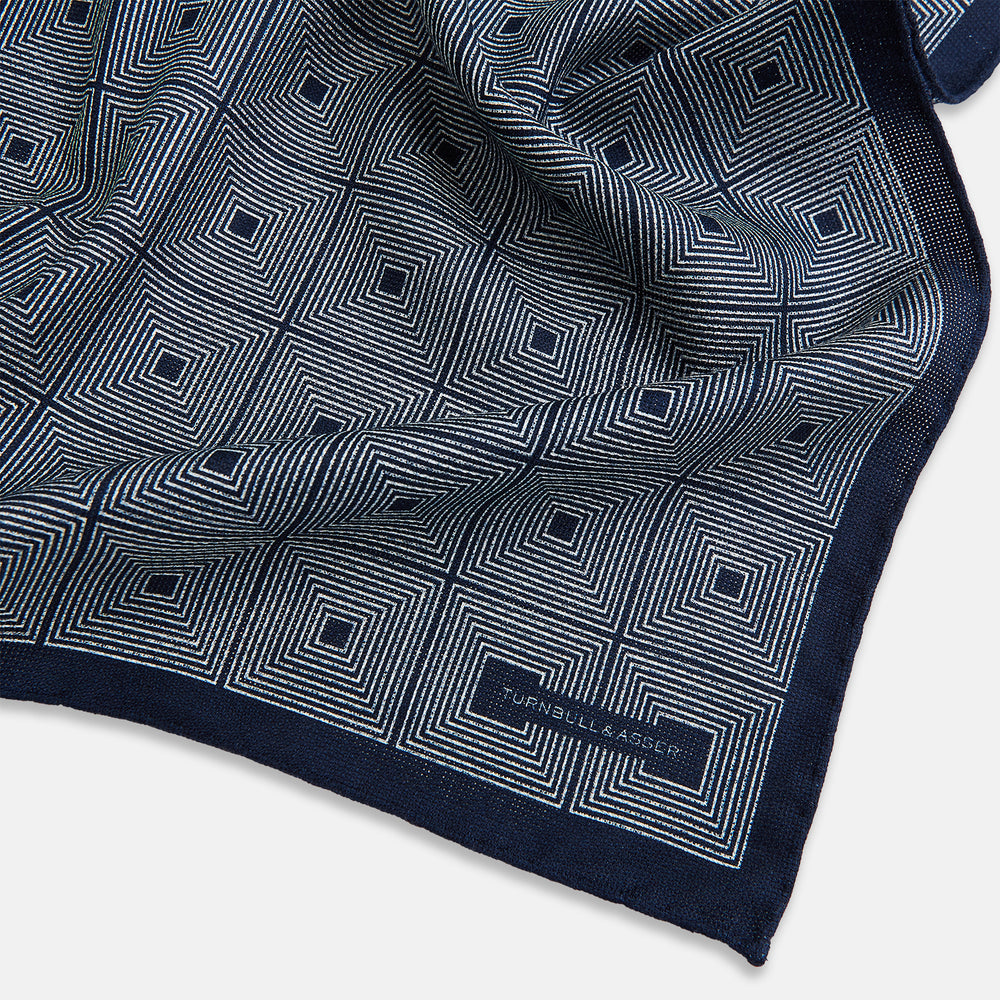 Introducing the Geometric Navy Tile Pocket Square in Silk by Turnbull & Asser UK—a folded navy and white accessory showcasing a double-sided design, with one side featuring concentric squares that form a striking geometric pattern and the other adorned with an elegant small dot motif.