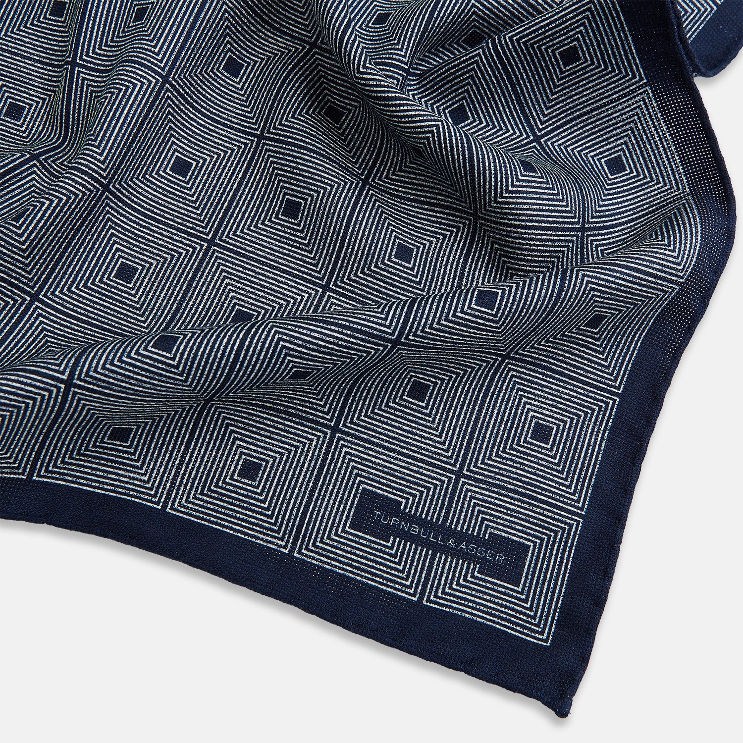 View 3 of product Introducing the Geometric Navy Tile Pocket Square in Silk by Turnbull & Asser UK—a folded navy and white accessory showcasing a double-sided design, with one side featuring concentric squares that form a striking geometric pattern and the other adorned with an elegant small dot motif.