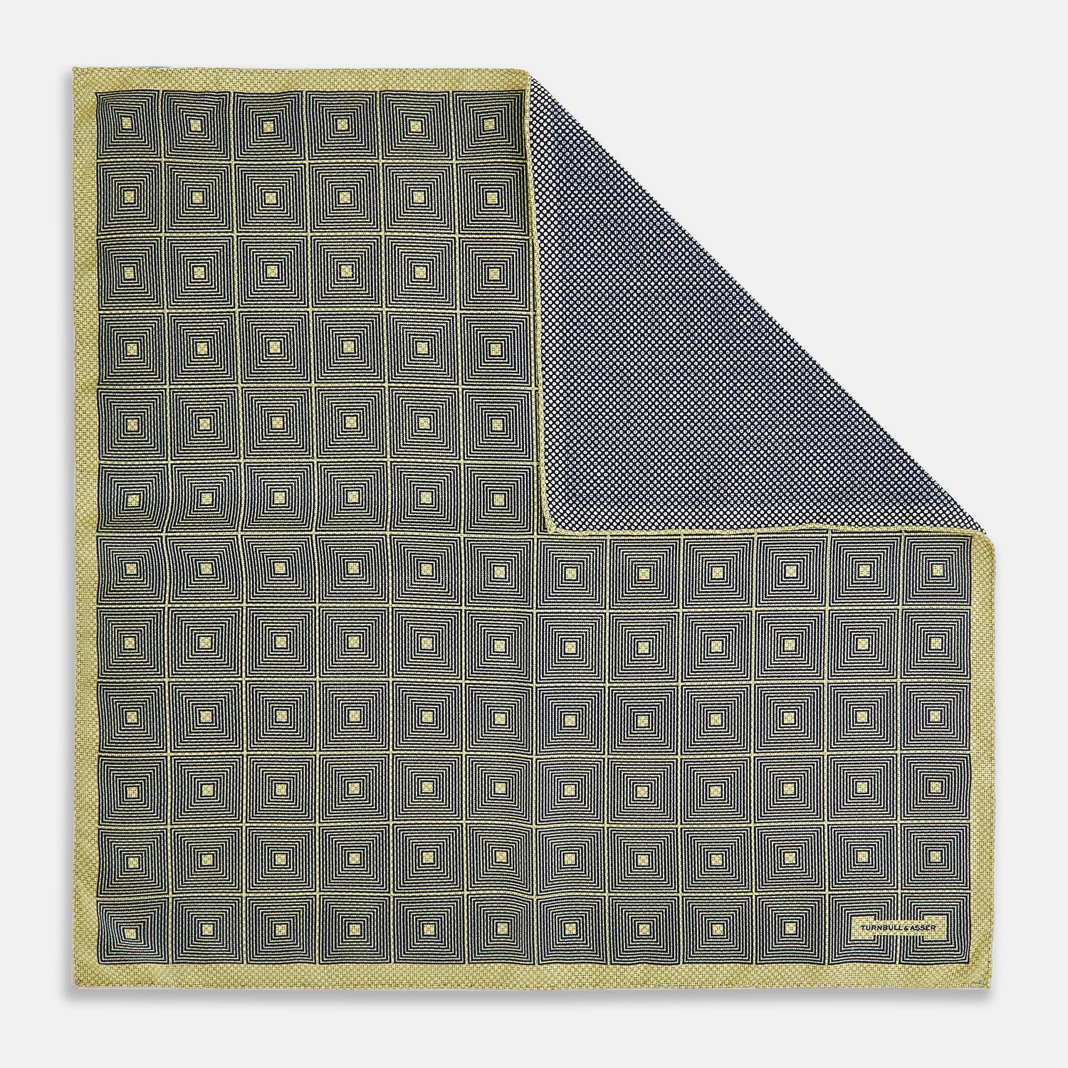 View 1 of product The Turnbull & Asser UK Geometric Yellow Tile Pocket Square in Silk features a sophisticated geometric square pattern in gold and navy, accented with subtle concentric squares and a discreet label stitched into the bottom right corner. Its double-sided design is highlighted by a folded corner that reveals a contrasting dotted navy and white underside.