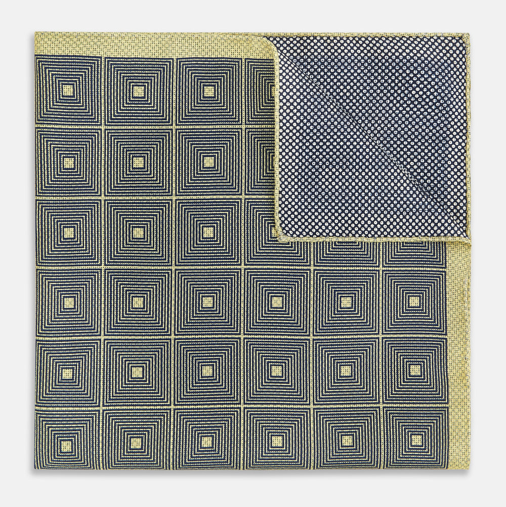 The Turnbull & Asser UK Geometric Yellow Tile Pocket Square in Silk features a sophisticated geometric square pattern in gold and navy, accented with subtle concentric squares and a discreet label stitched into the bottom right corner. Its double-sided design is highlighted by a folded corner that reveals a contrasting dotted navy and white underside.