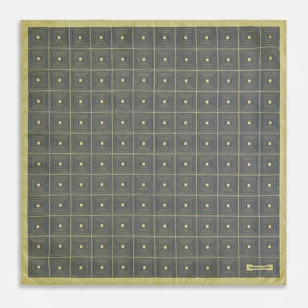 The Turnbull & Asser UK Geometric Yellow Tile Pocket Square in Silk features a sophisticated geometric square pattern in gold and navy, accented with subtle concentric squares and a discreet label stitched into the bottom right corner. Its double-sided design is highlighted by a folded corner that reveals a contrasting dotted navy and white underside.