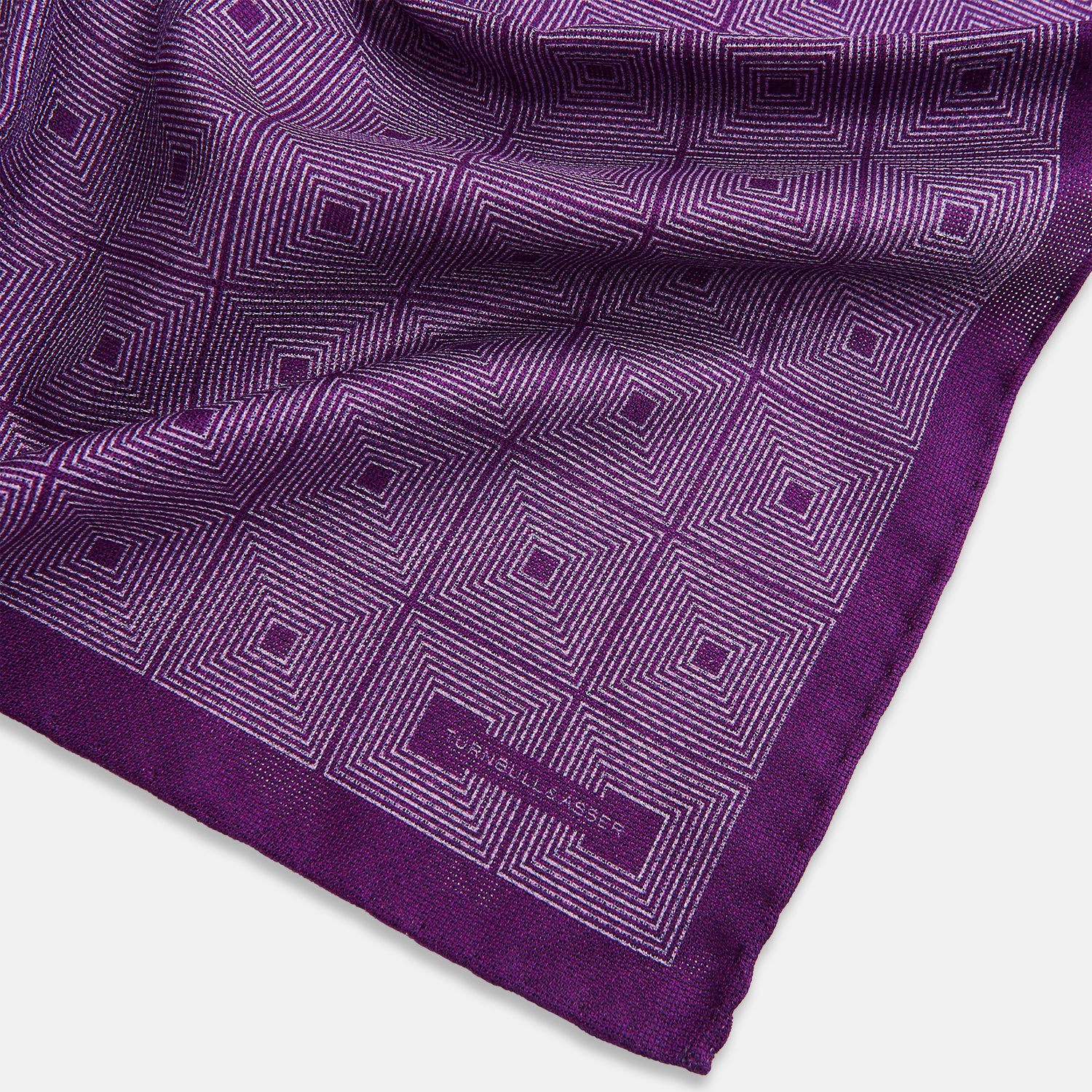 View 3 of product The Turnbull & Asser UK Geometric Purple Tile Pocket Square in Silk features a vibrant purple hue adorned with concentric square motifs. One corner is folded to reveal its double-sided design, showcasing a contrasting black-and-white dotted underside.